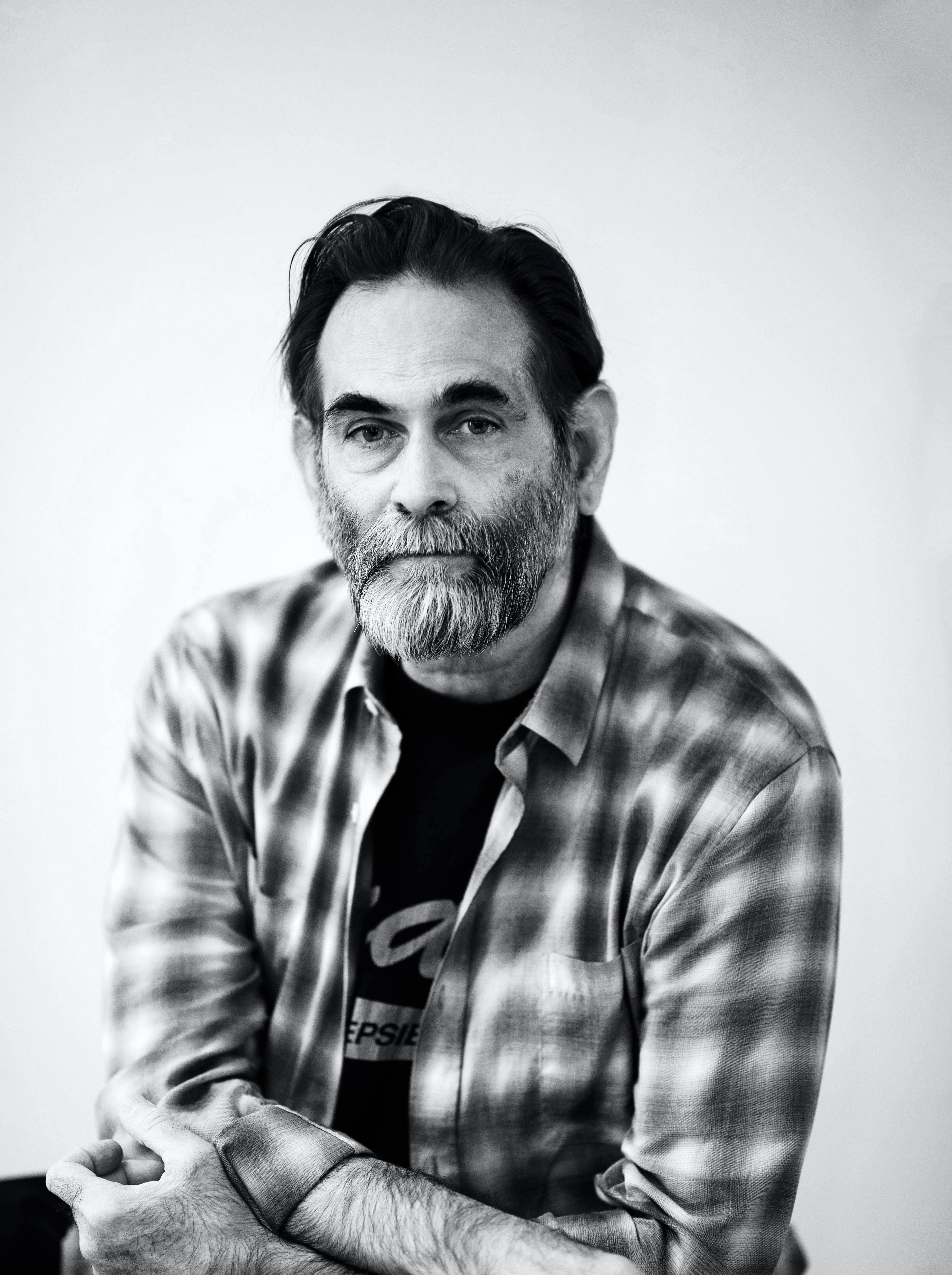 A black and white photo of Avram, a white person, sitting down looking straight at the camera. He has black hair, a salt and pepper beard and is wearing a flannel shirt over a black t-shirt.