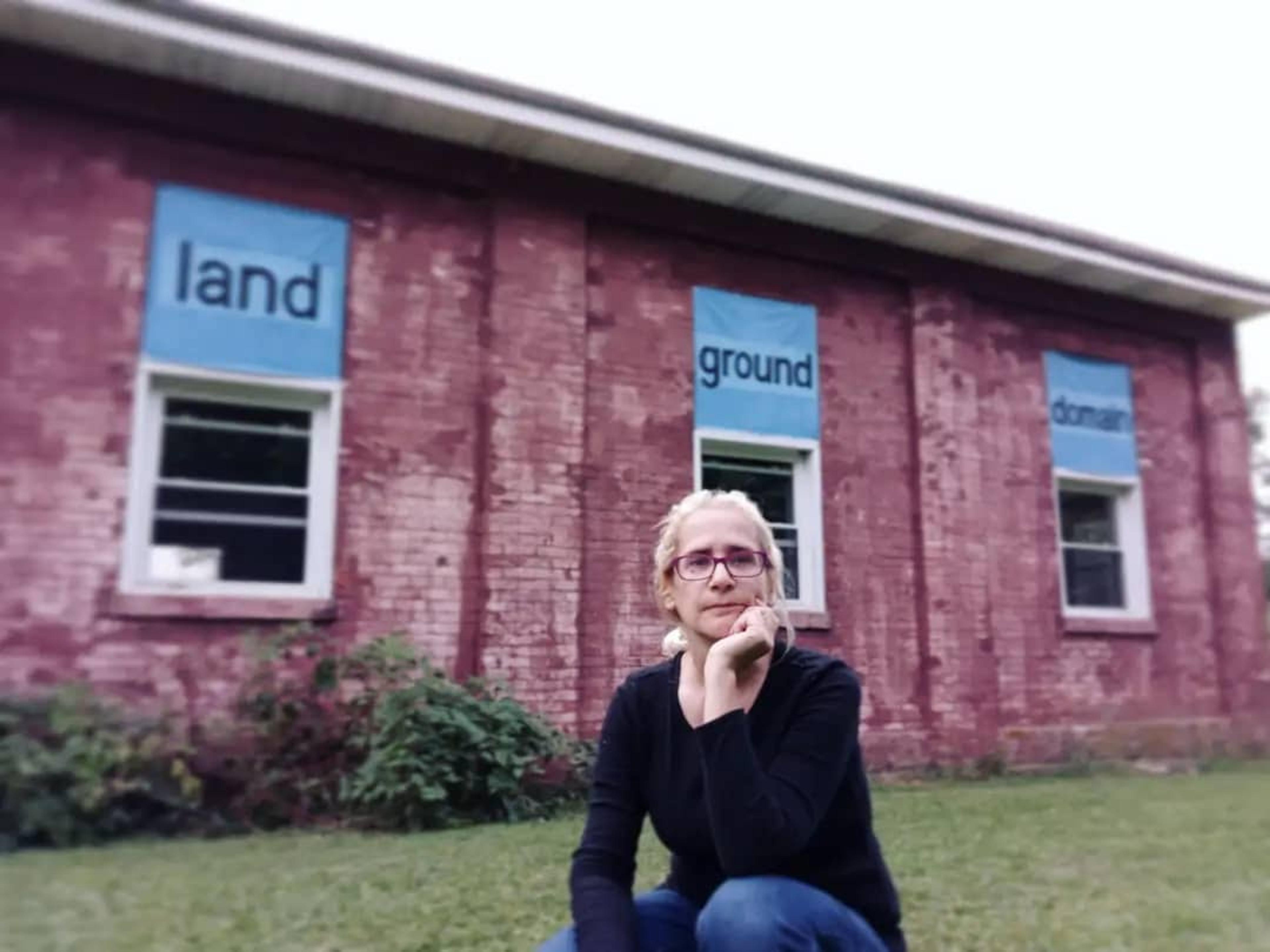 A photo of Mona, a blond-haired middle-eastern woman wearing eyeglasses, a black top and blue jeans. Mona is crouching on the grass in front of a red brick building. On the building are 3 square banners painted sky blue with dark brown lettering.