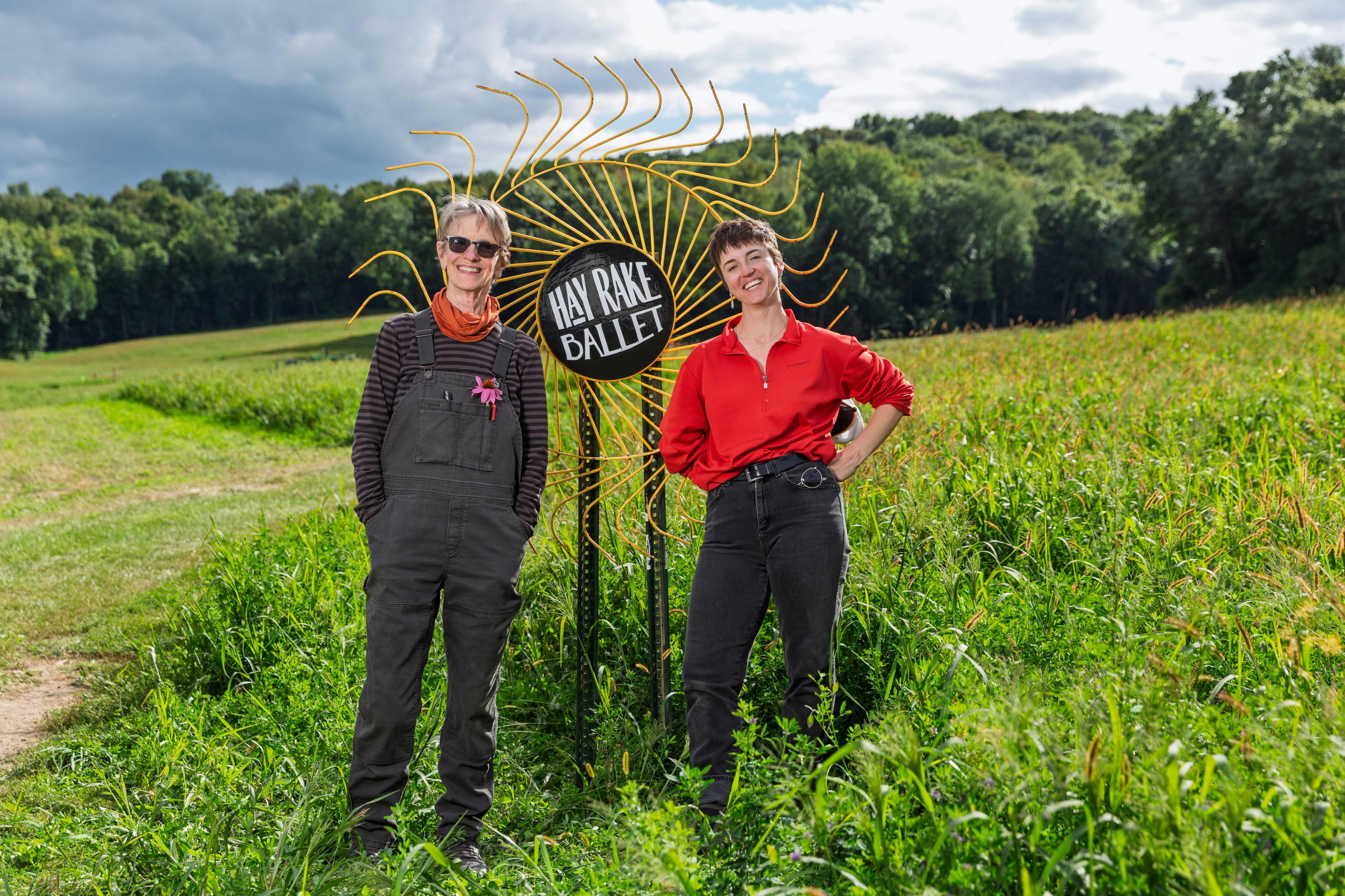Two people stand in a green field next to a sign that says Hay Rake Ballet.