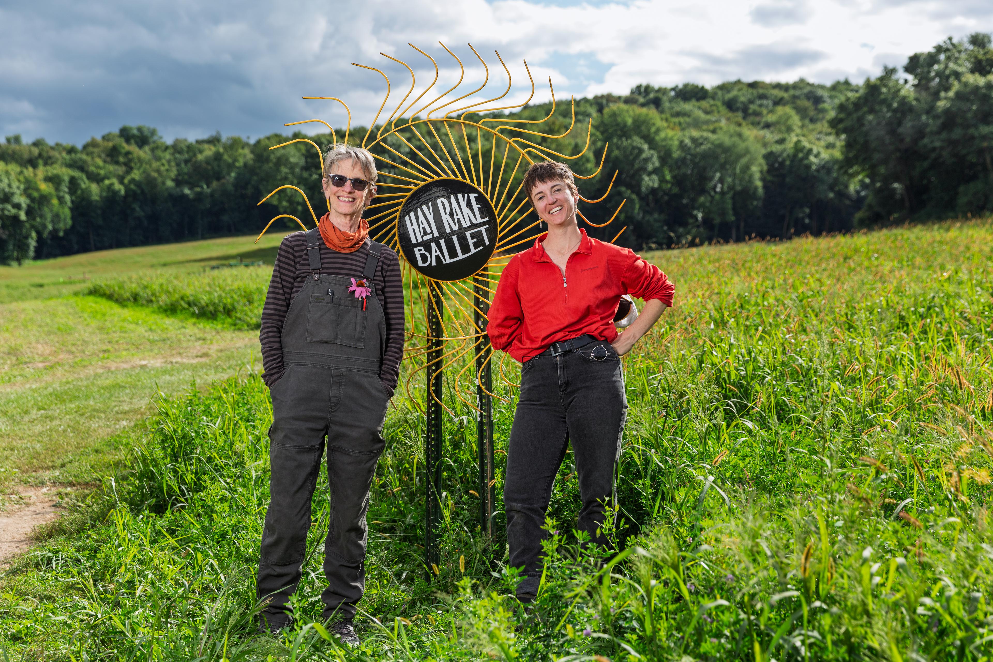 Two people stand in a green field next to a sign that says Hay Rake Ballet.