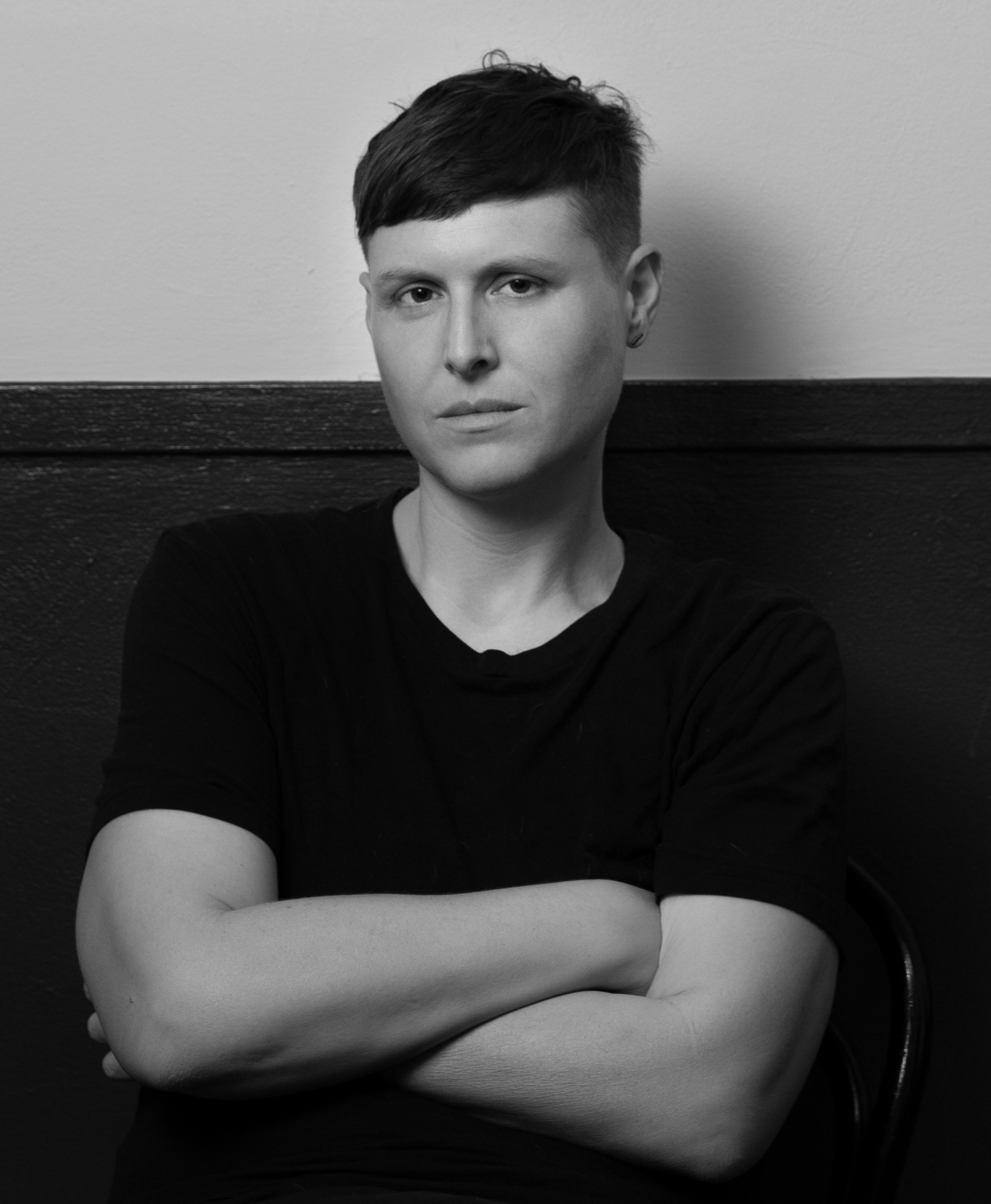 A black and white photo of LJ Roberts. LJ, a White person with short dark hair, sits with their arms crossed, looking intently at the camera. They are wearing a black t-shirt. 