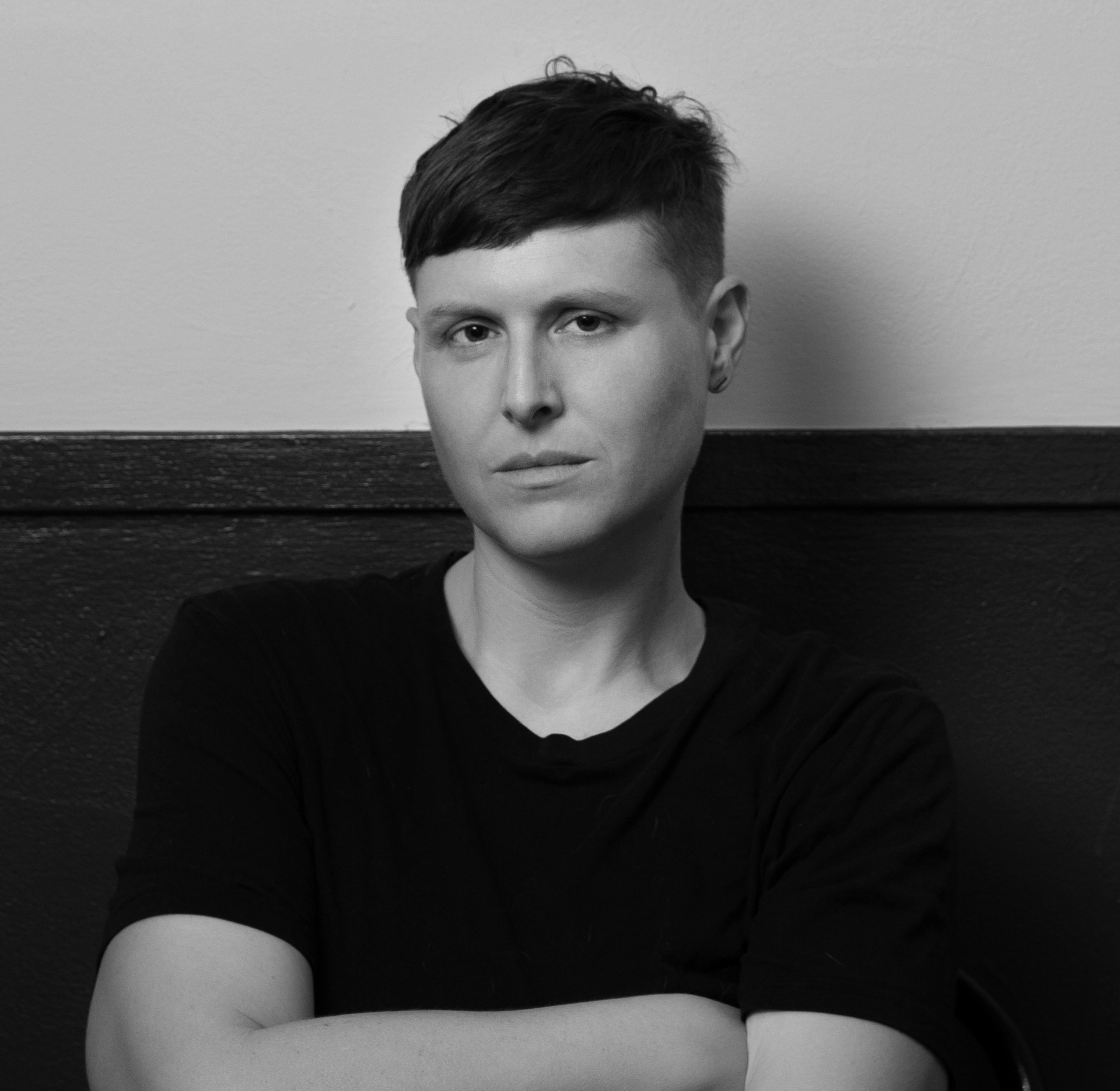 A black and white photo of LJ Roberts. LJ, a White person with short dark hair, sits with their arms crossed, looking intently at the camera. They are wearing a black t-shirt.