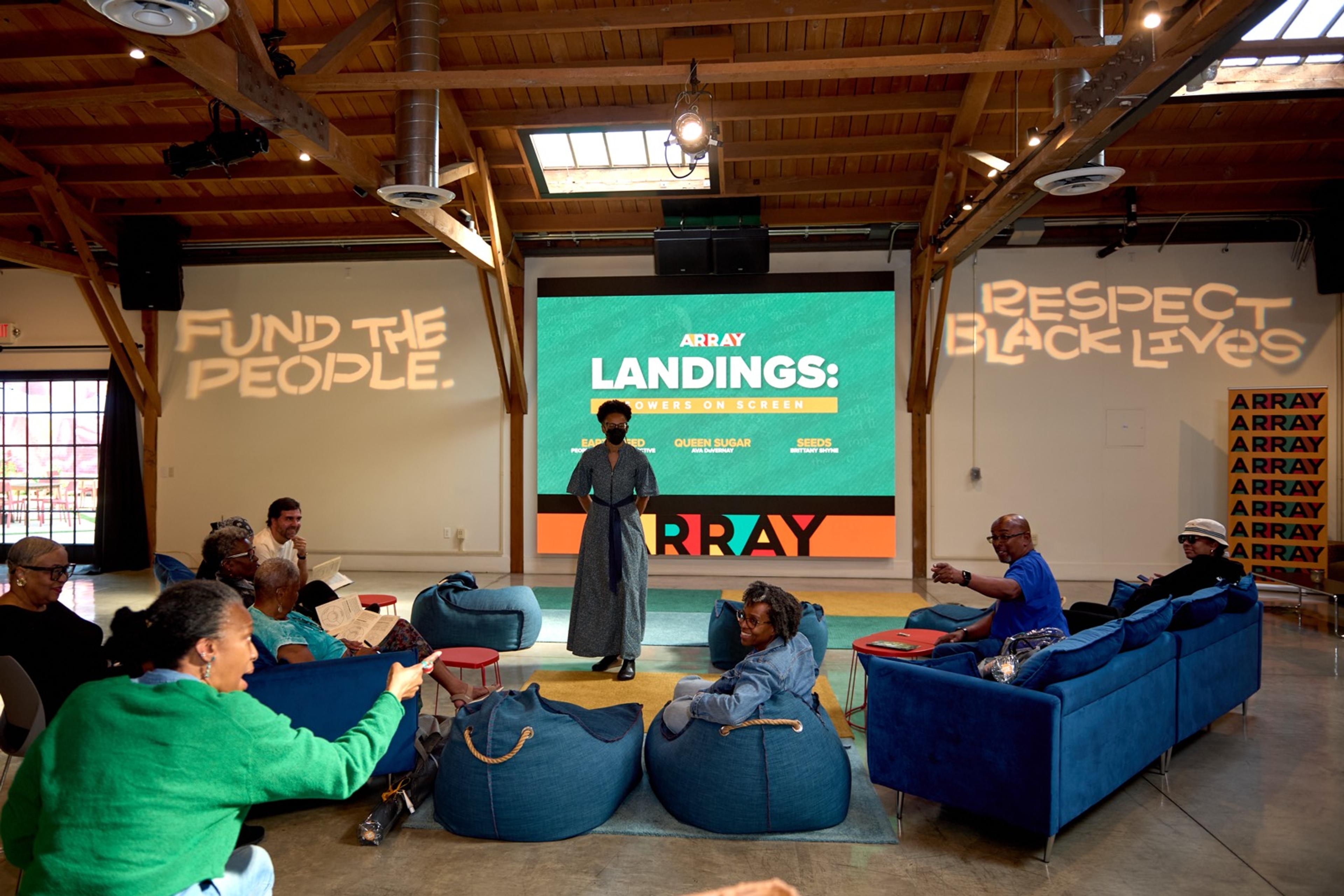 Jocelyn stands in front of a screen, introducing the EARTH SEED film. The audience sits around her on bean bag chairs and couches.