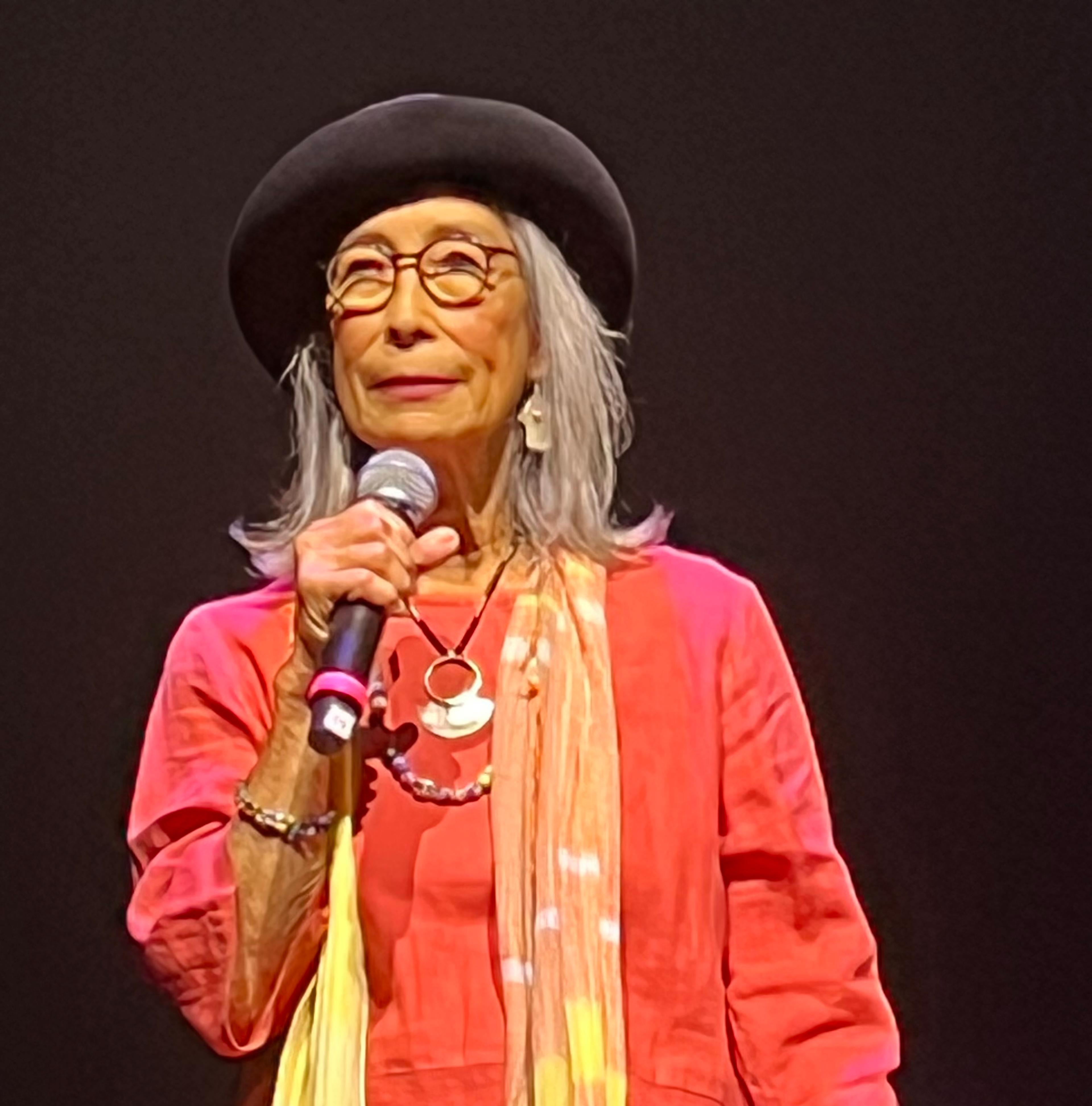 A Japanese American woman holds a microphone and stands on a dark stage. She wears bright orange flowy clothes and a yellow and orange scarf, round glasses, a beret, and silver jewelry.