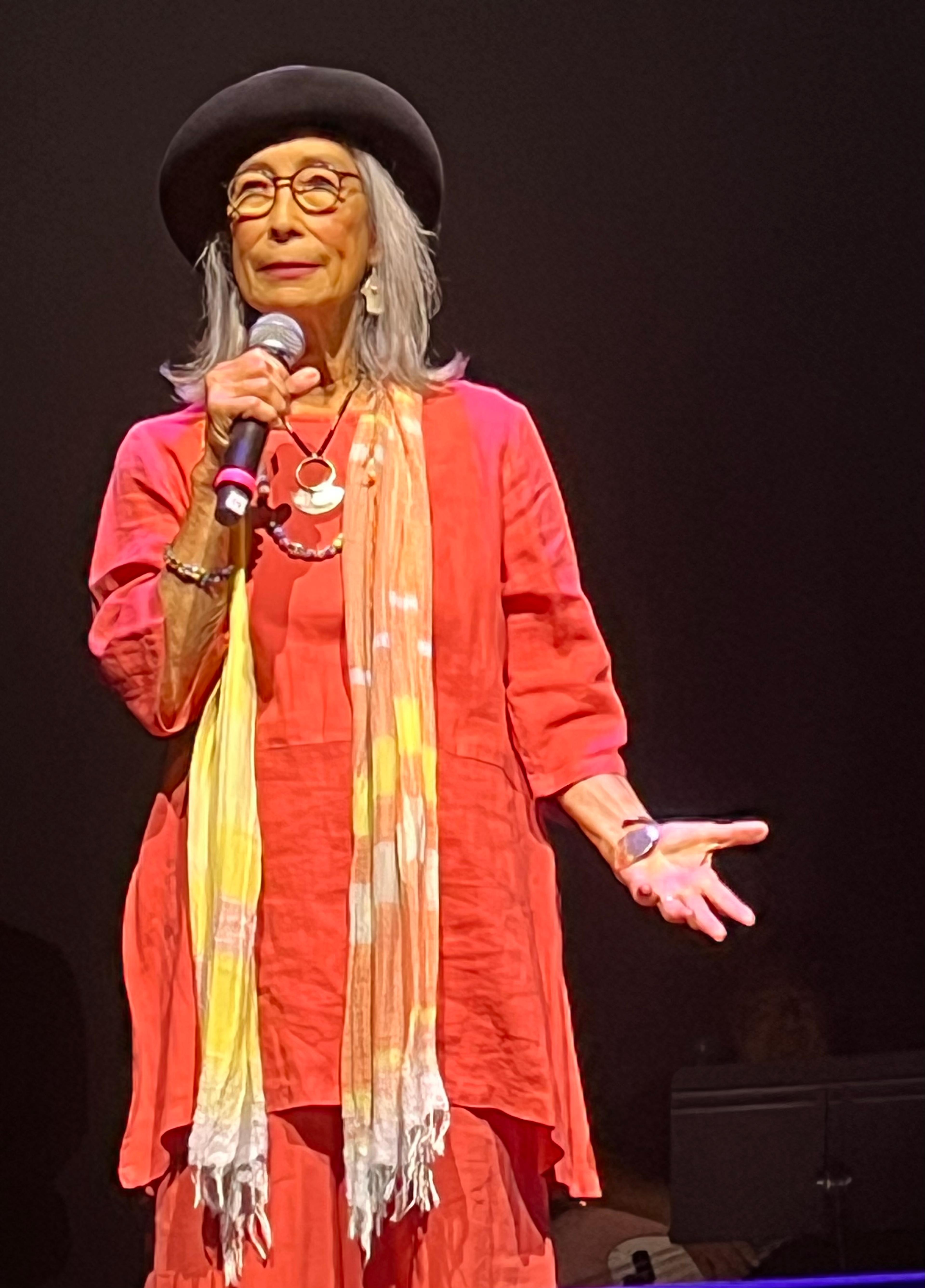 A Japanese American woman holds a microphone and stands on a dark stage. She wears bright orange flowy clothes and a yellow and orange scarf, round glasses, a beret, and silver jewelry.