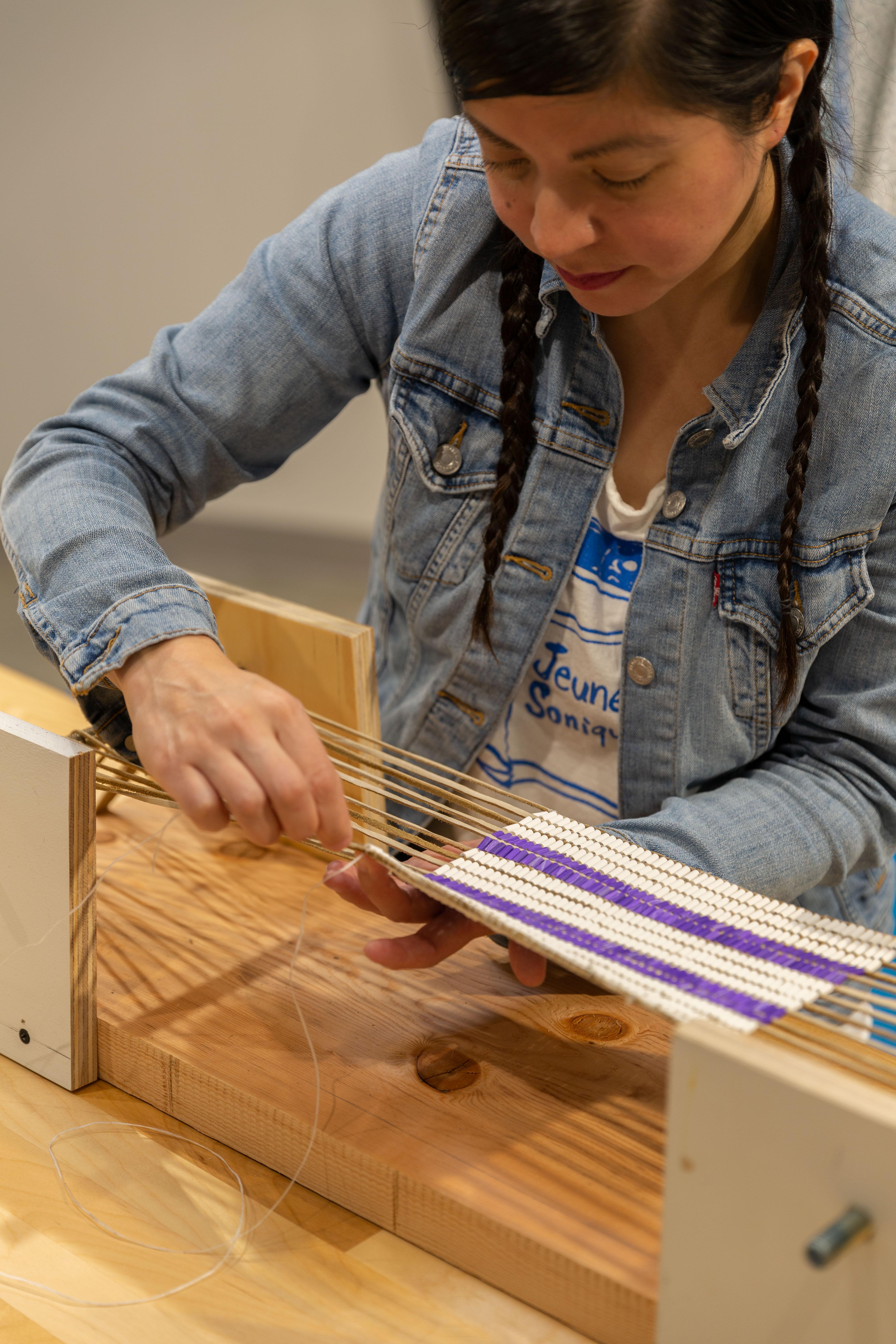 An Indigenous woman with braids wearing a jean jacket is making a two row wapum belt on a homemade loom.