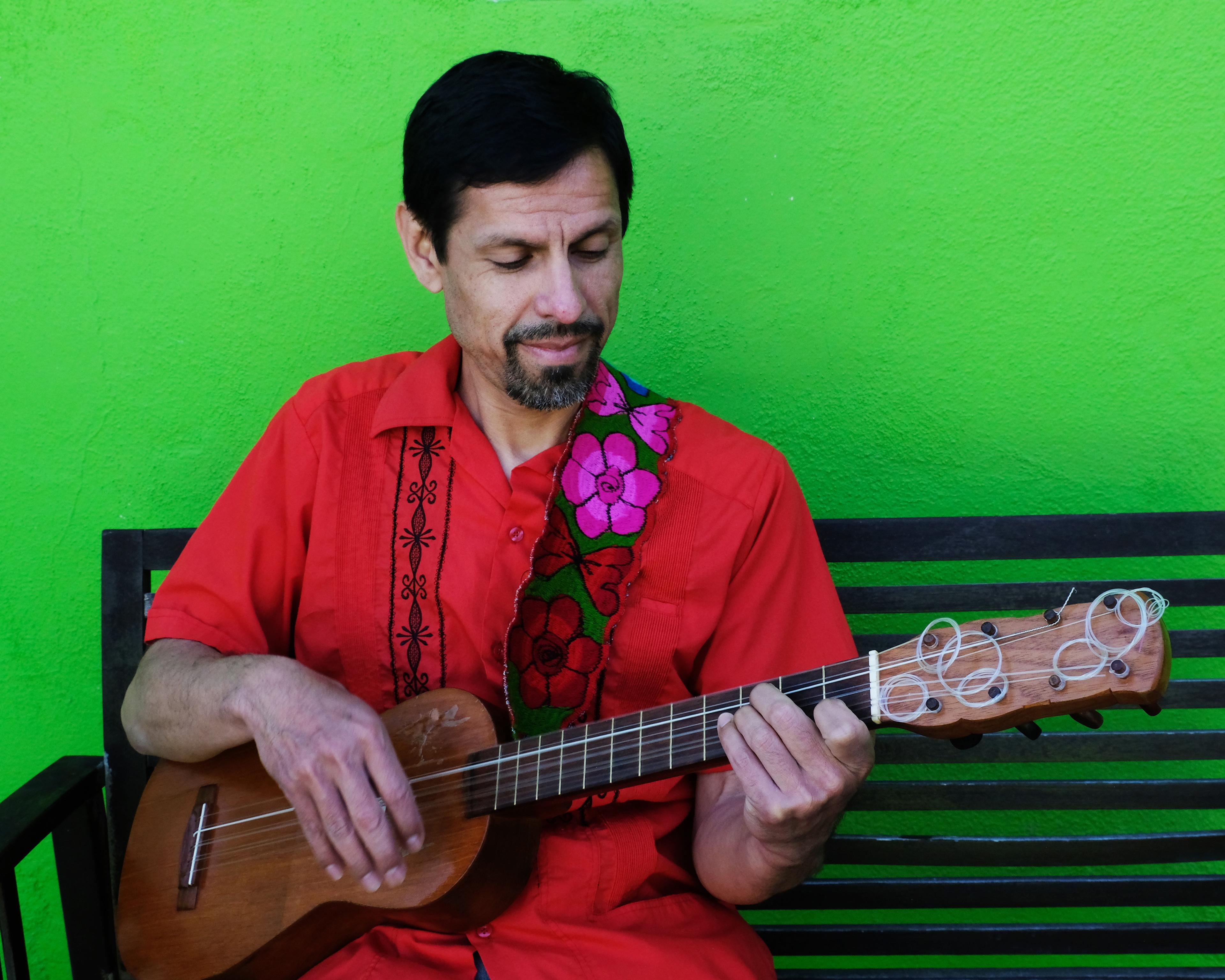 A man in a bright red shirt plays a jarana, a guitar-shaped fretted stringed instrument. The wall behind him is bright lime green.