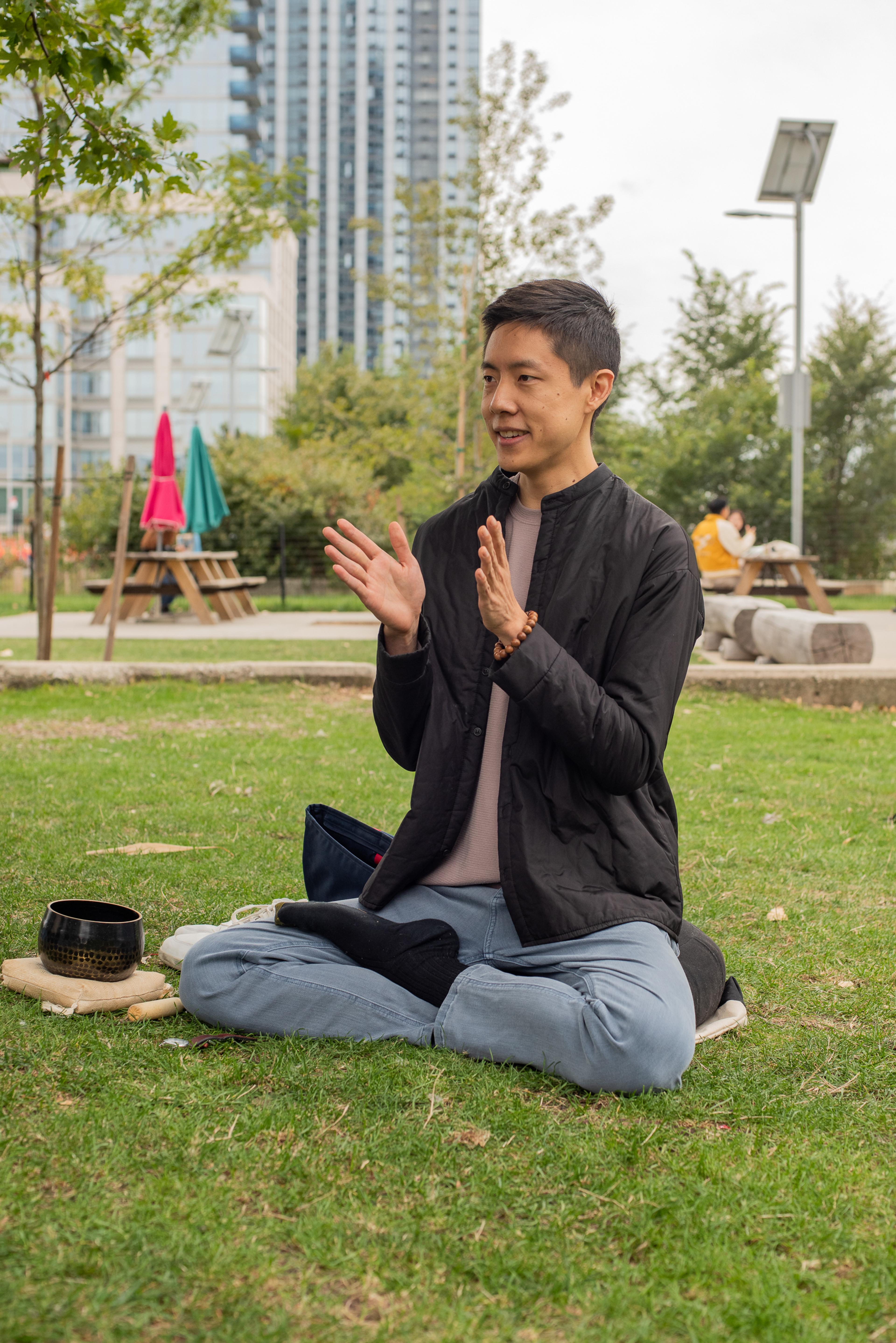 Ryan sits cross-legged on the lawn of a city park. His hands are mid-clap and to his right is a singing bowl on a small pillow.