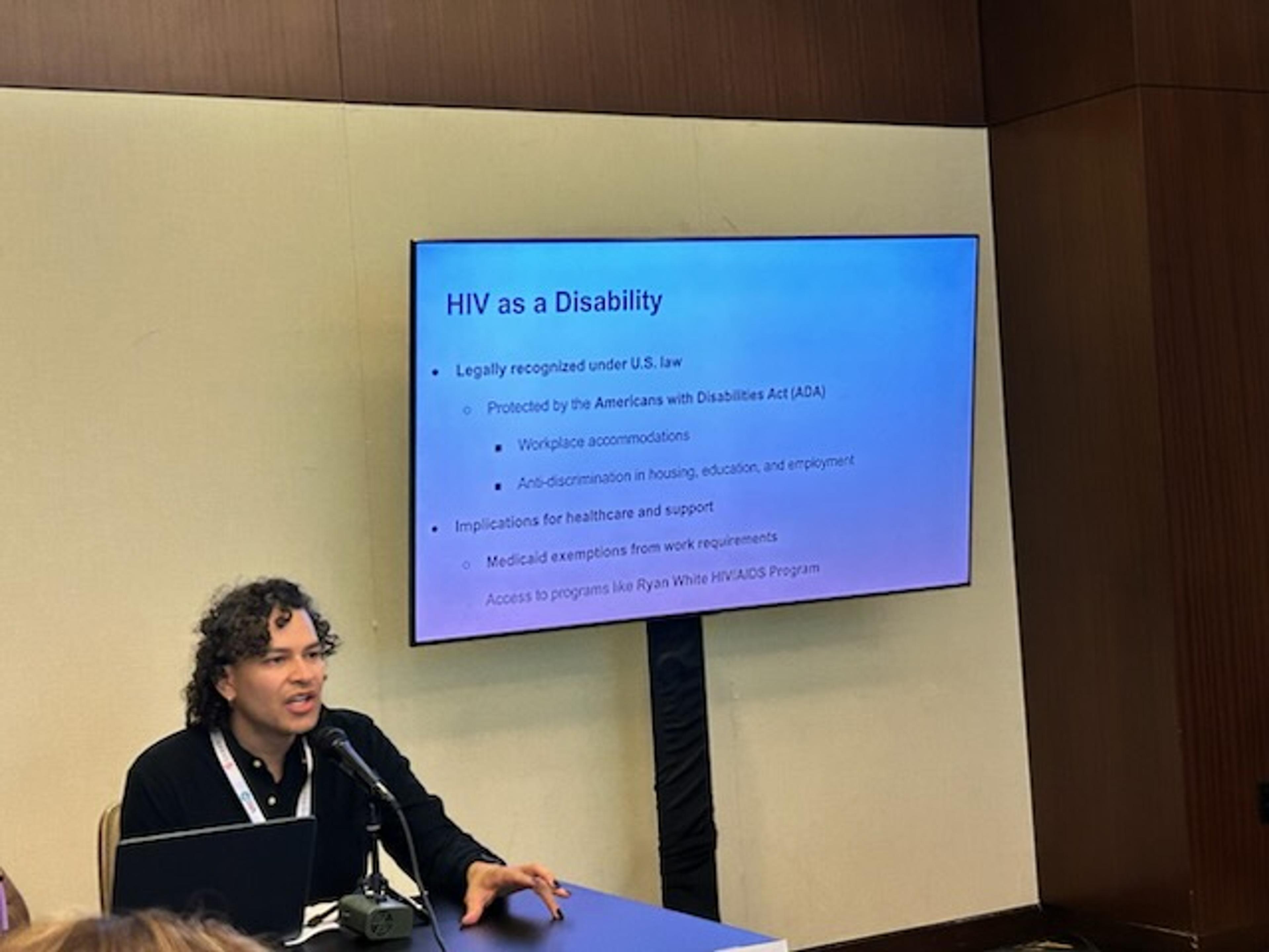 Photo of a light-skinned Latine genderqueer person seated at a table and speaking into a microphone while gesturing with one hand. They have short, dark, curly hair and are wearing a dark long-sleeve top. They are wearing a conference lanyard and using a microphone. The background shows a conference setting with wood-paneled walls and a presentation slide on a screen behind them titled “HIV as a Disability.”
