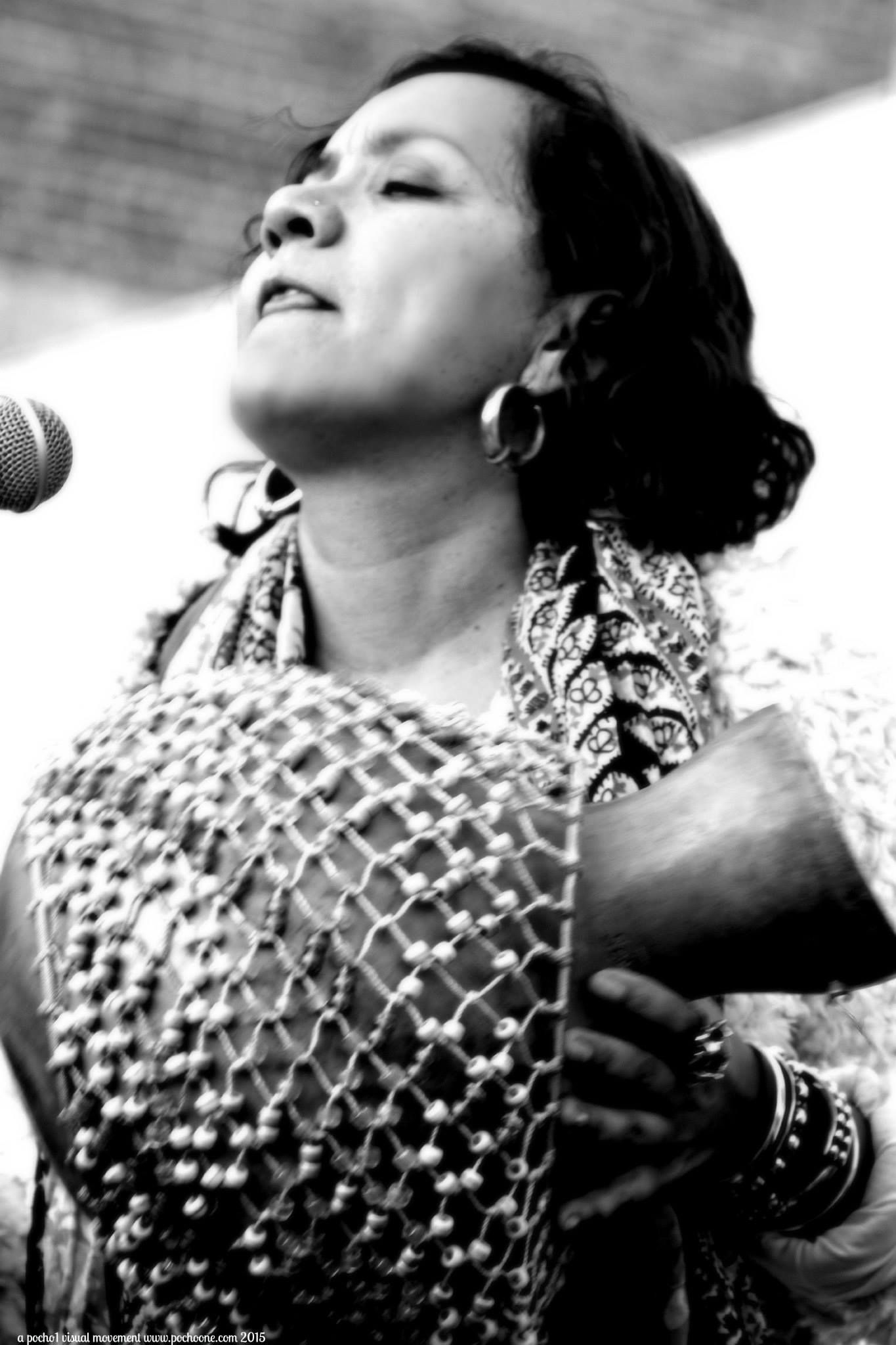 Black and white image of a brown woman holding a percussion instrument and looking up in delight as she plays joyfully.