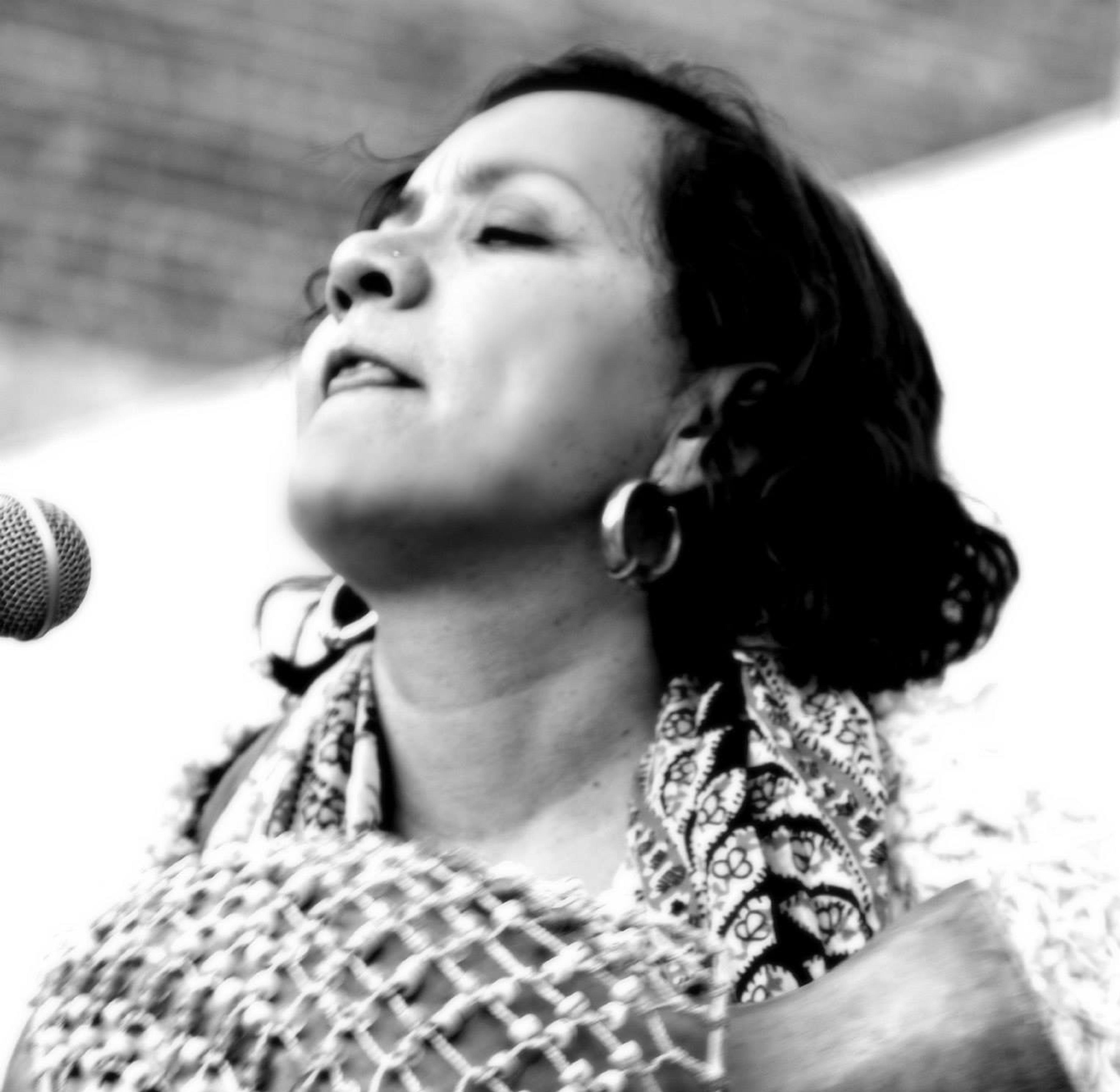 Black and white image of a brown woman holding a percussion instrument and looking up in delight as she plays joyfully.