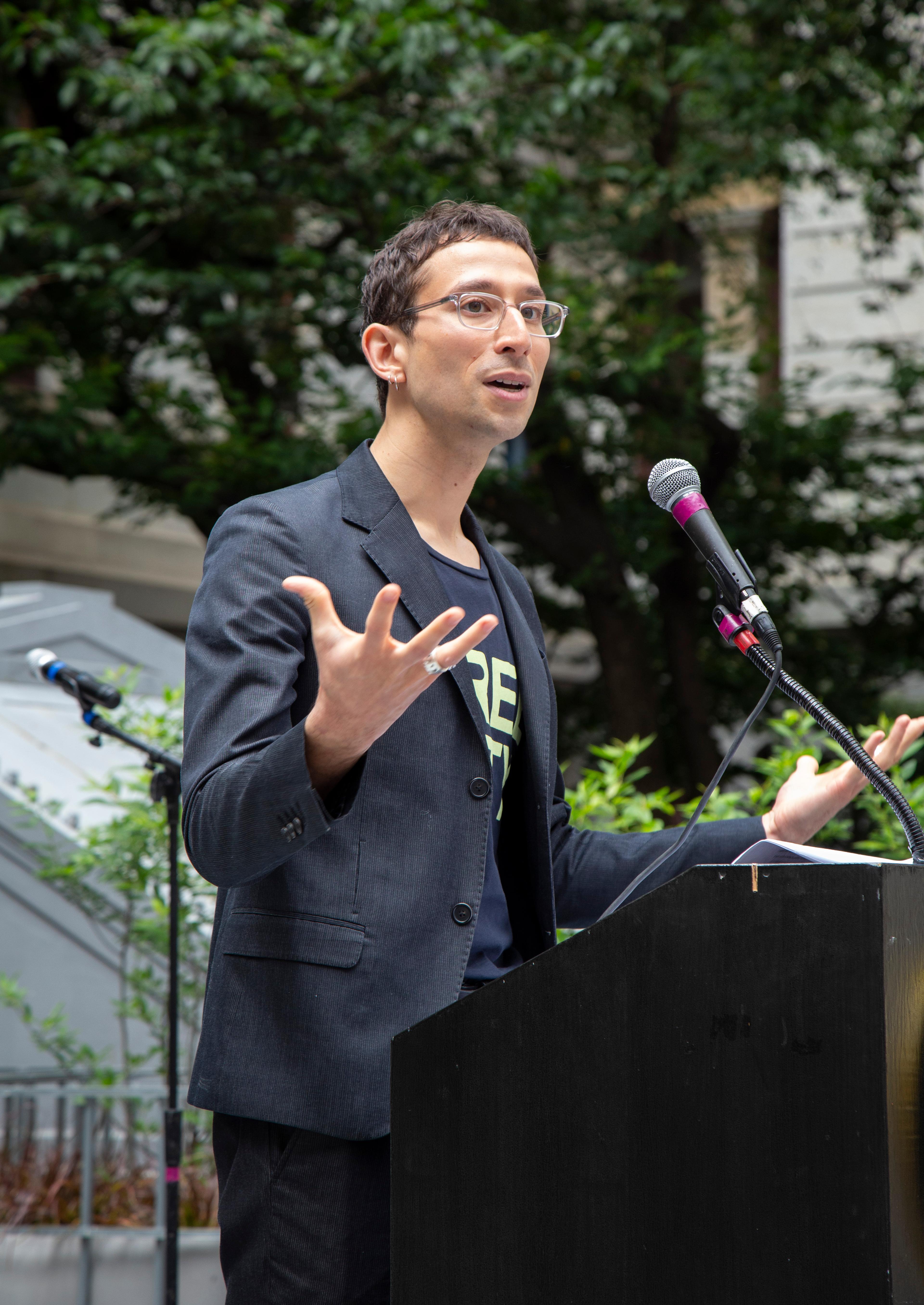 Emilio wears clear glasses and a blazer and stands at a podium, gesturing expressively.