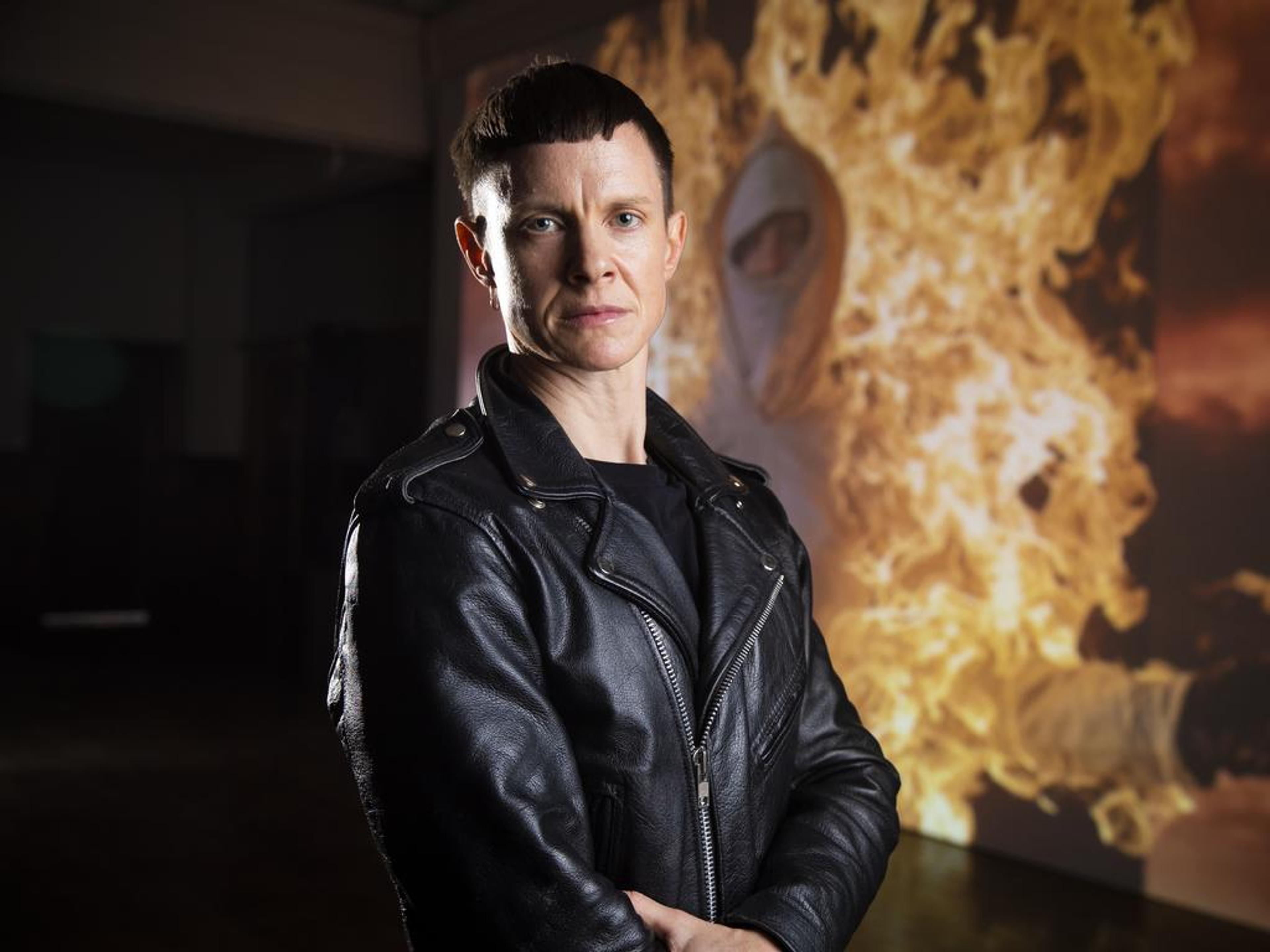 A photo of Cassils, a white person standing in front of a large projection of an artwork that depicts a masked figured engulfed in flames. Cassils is looking directly at the camera, they have short brown hair and are wearing a black leather jacket.