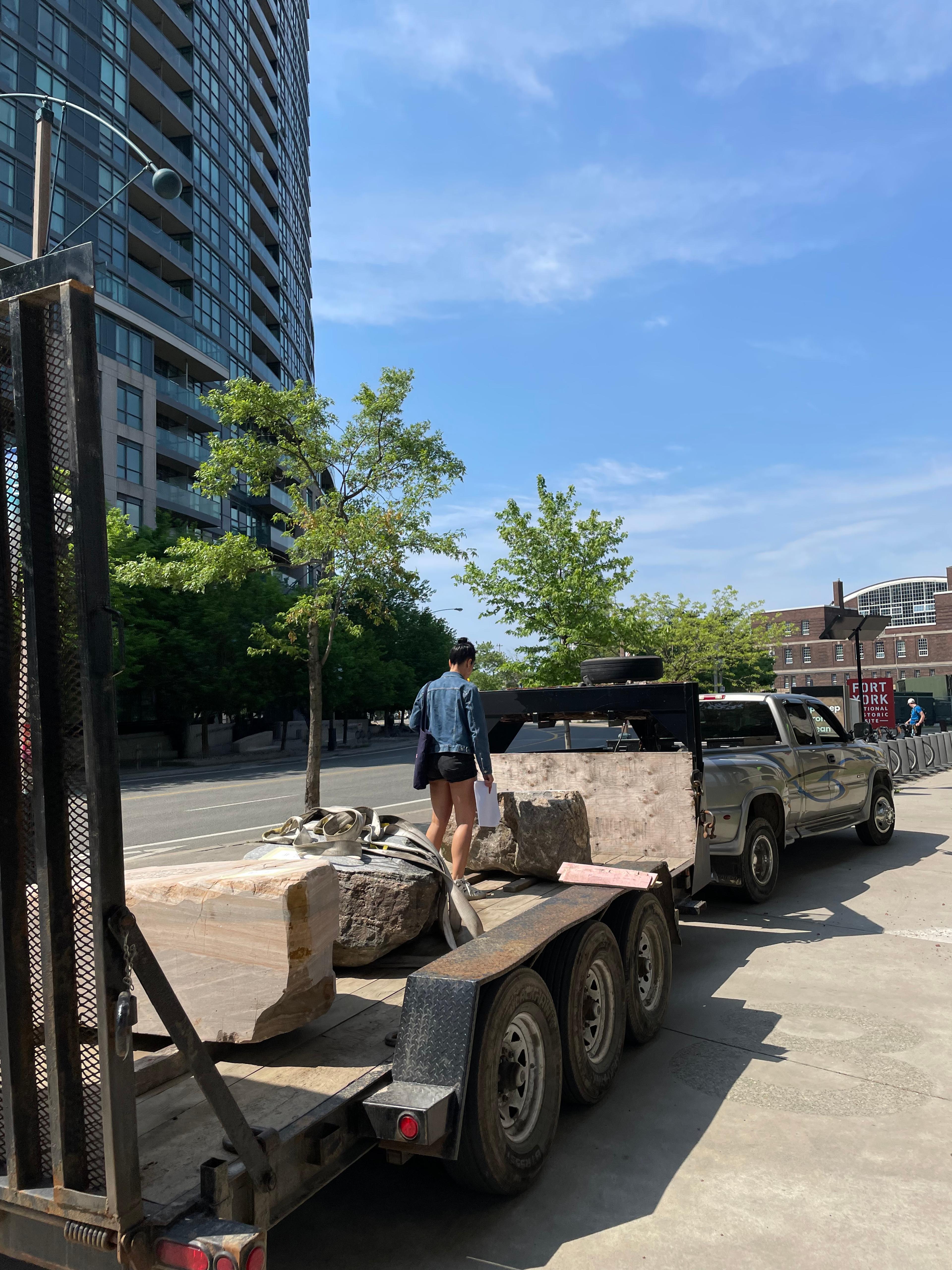 A photo of Chloë, a mixed-race Black American, with her back to the camera. She is wearing black denim shorts and a blue denim jacket. Her black hair is up in a bun.  She is standing on the back of an open-air flatbed truck.  There are three large stone bench sculptures on the back of the truck as well, and Chloë is looking down at one of them. The background includes a glass apartment tower to the left, blue sky with wispy white clouds above, several small and leafy trees, and a sign in the background reading "Fort York National Historic Site."