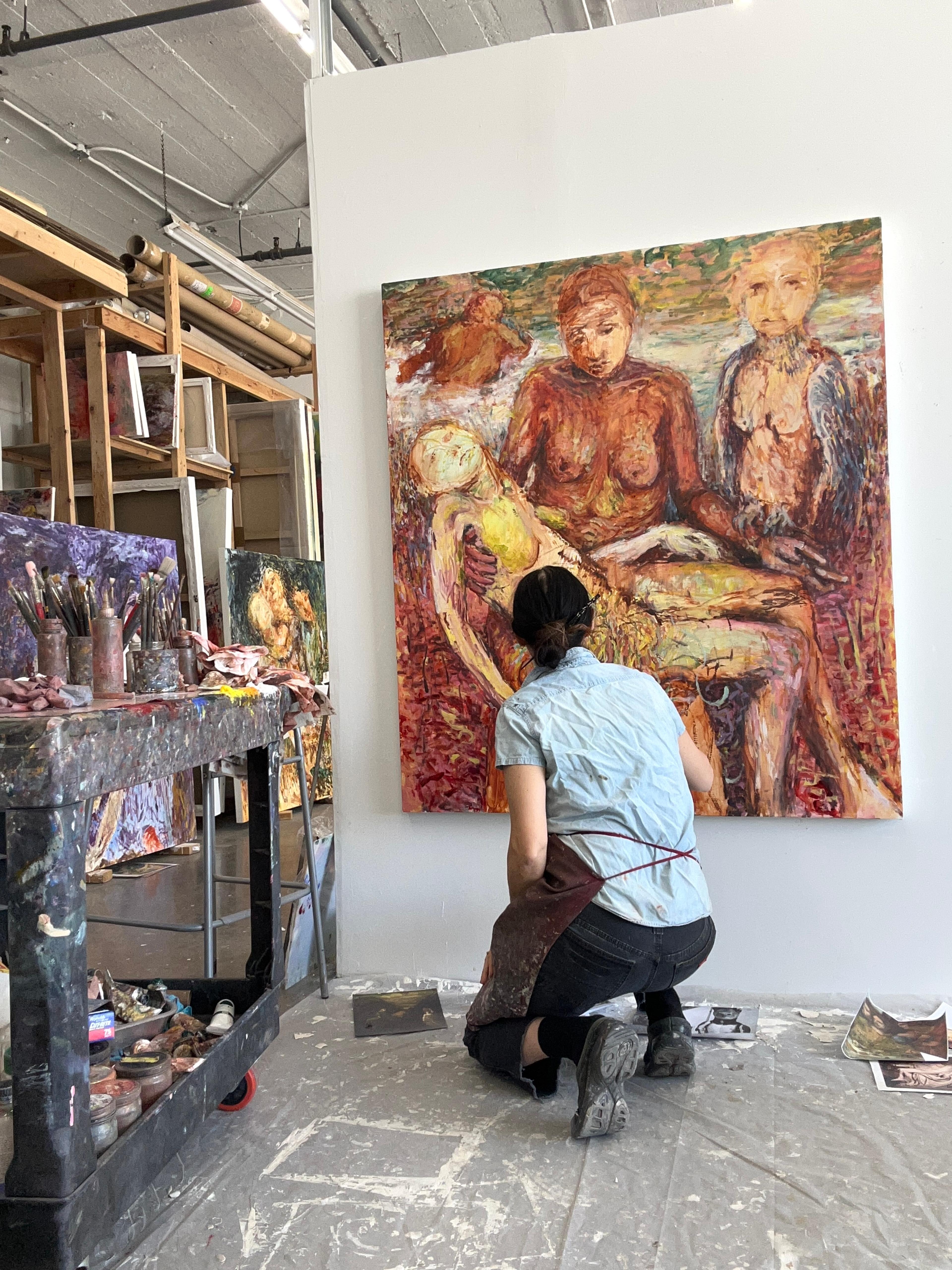 An artist is in their studio working on a painting. She is crouched down on one knee with her back facing the camera and is wearing a light blue shirt and black pants. There is a palette table with brushes on the left and other paintings and painting racks in the background.  
