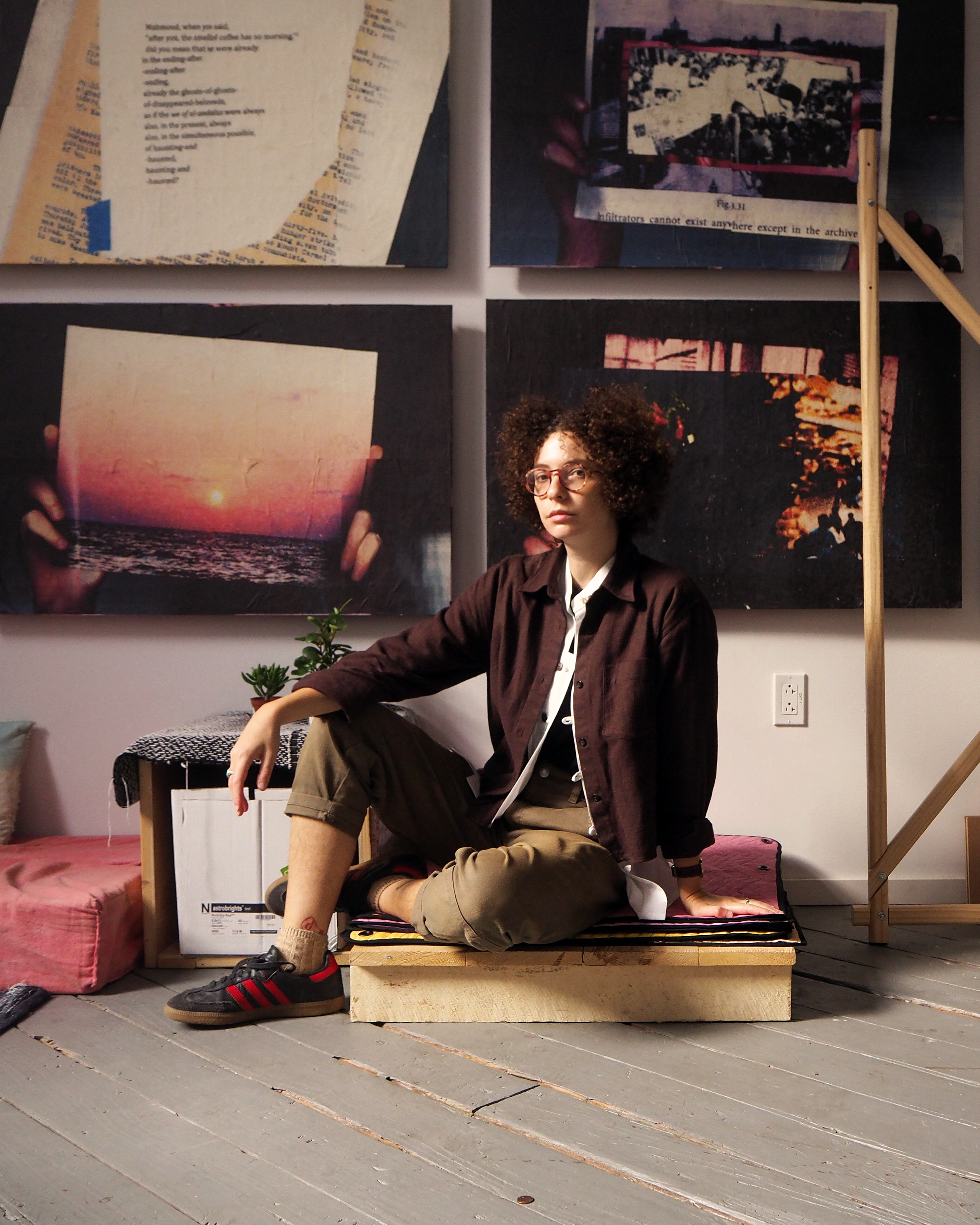 kimi malka hanauer sits on a wooden pallet in their studio. kimi is of sephardic and ashkenazi descent, has curly hair, glasses, and wears flannel a button up shirt. Their legs are folded, one beneath them, and the other placed on the floor facing the right. Behind them, a grid of artworks hang on the wall, featuring oversized scans of hands holding various materials. Most prominently, hands hold up an image of a sun setting over a body of water. The floor is grey and the afternoon sun lights kimi's face from the left.