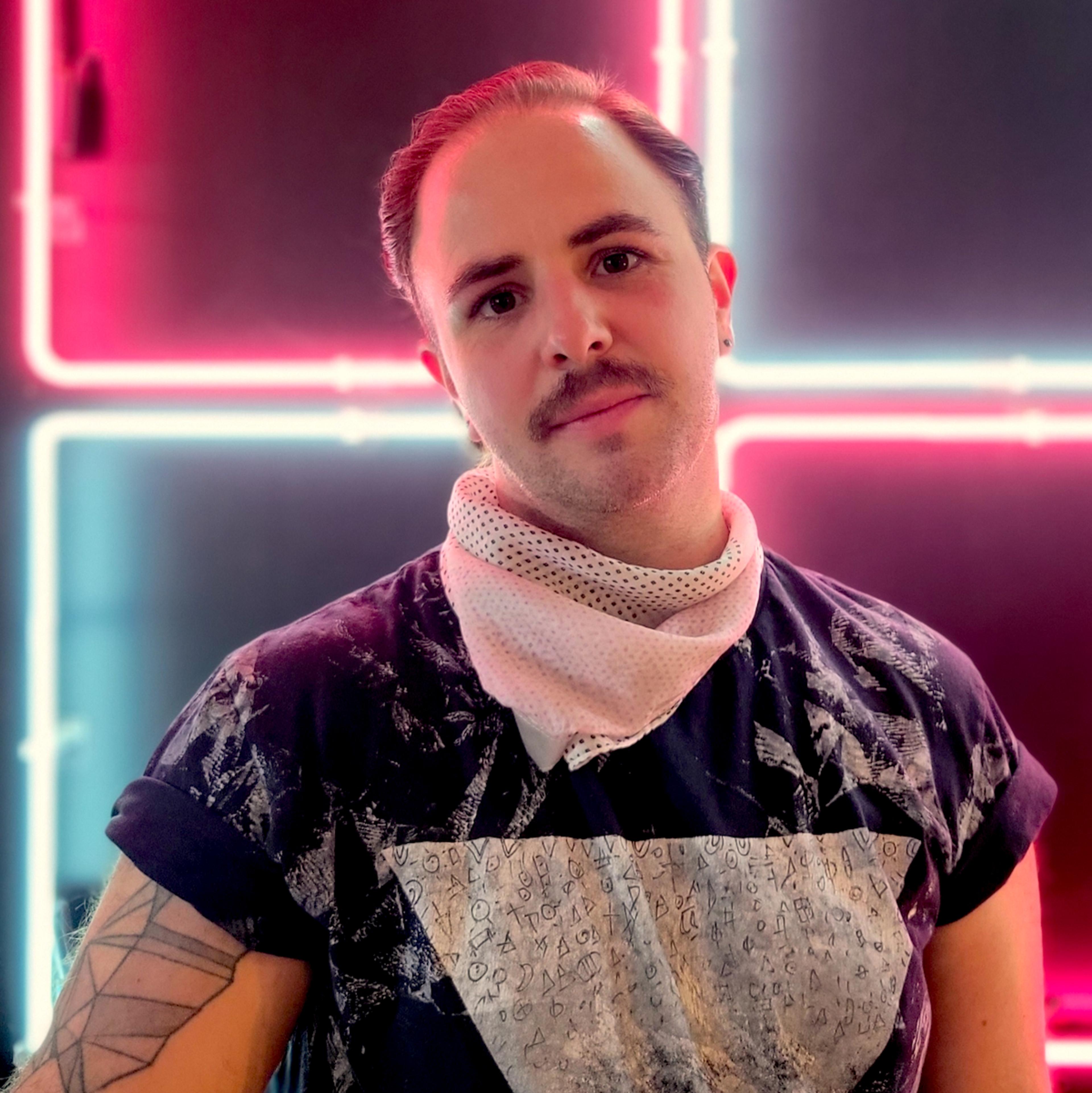 Anders, a white effeminate person standing in front of an illuminated white and hot pink neon sign, has brown hair and a mustache and wears a black and white t-shirt and a pink neckerchief.