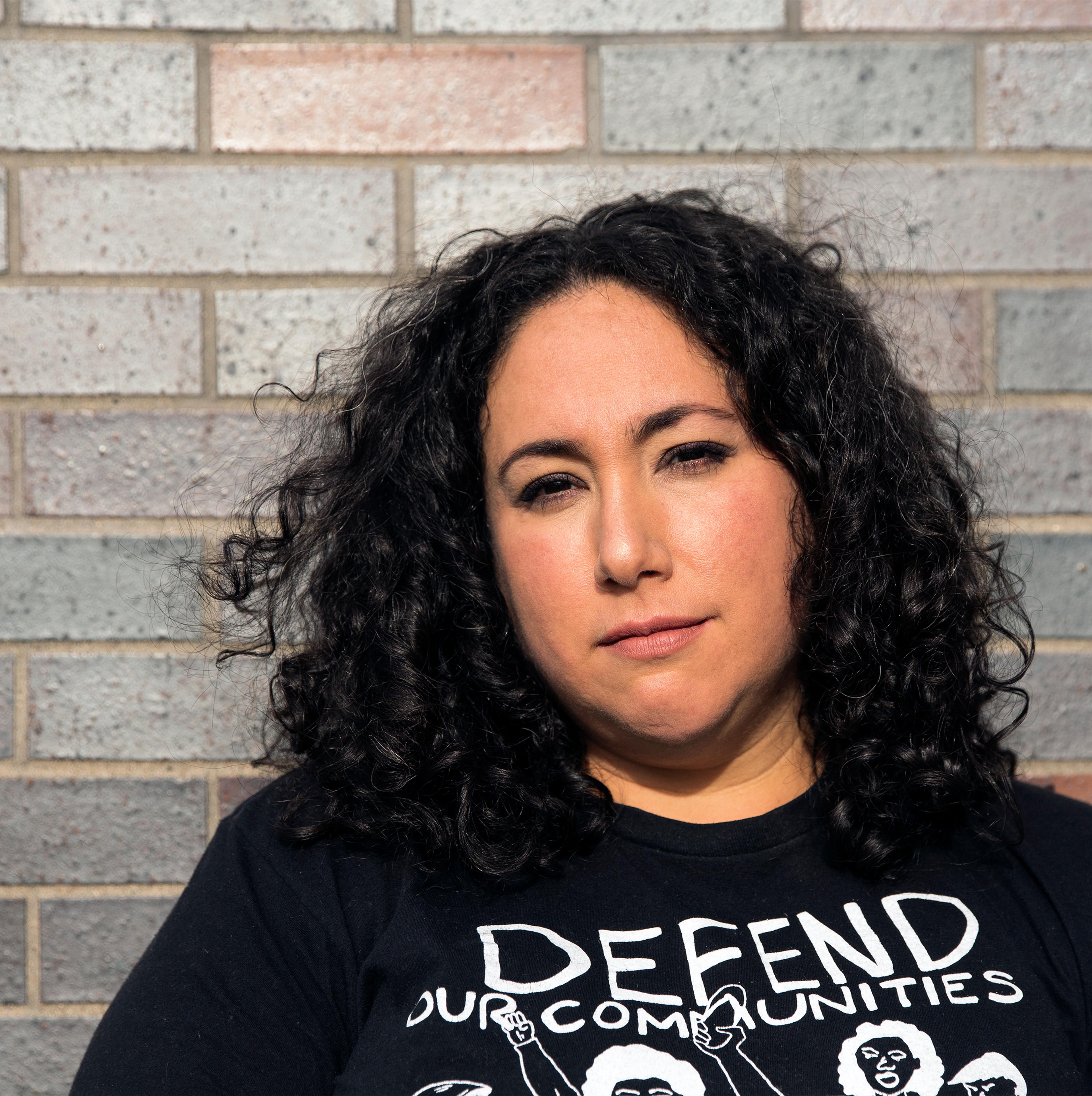 A photo of Risa, a medium-skinned brown woman with dark, shoulder-length curly hair stands in front of a brick wall. She wears a black t-shirt designed by Chicago-based artist  Monica Trinidad with the words “defend our communities” written in white lettering. The outline of raised fists are drawn under the letters. 