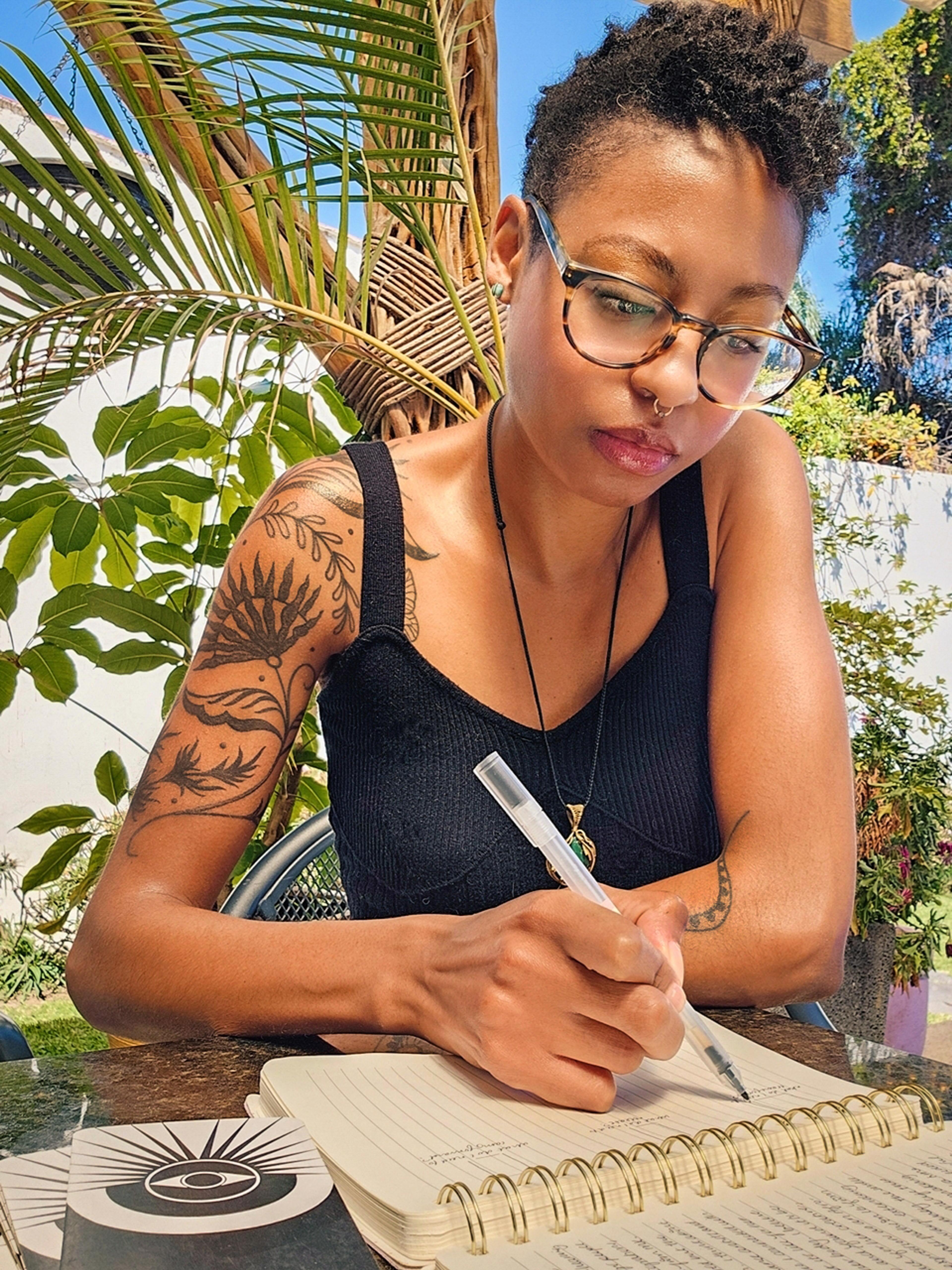 A photo of Denise Shanté, a black woman, sitting at a table writing in a garden. She is wearing glasses, has a septum hoop, and a sleeve of tattoos on her right arm. She is wearing a black tank top.