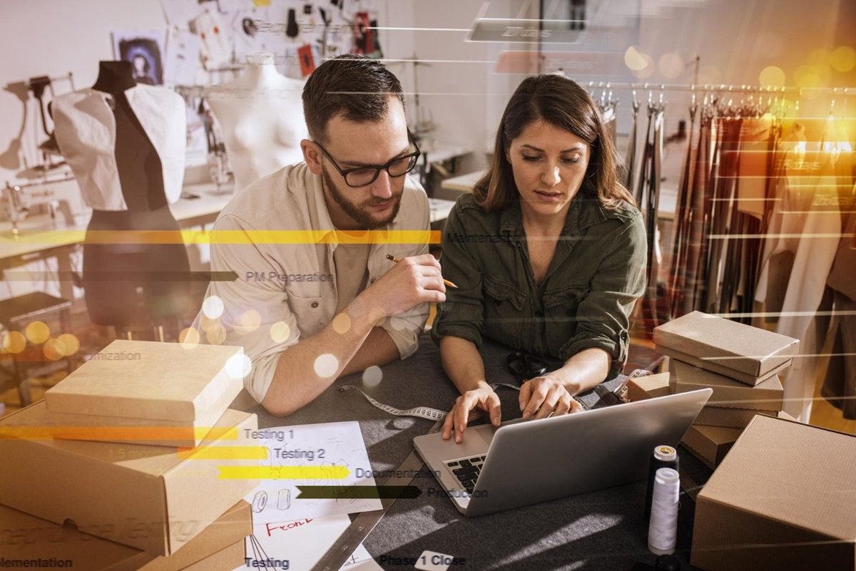 Pareja de emprendedores revisa una laptop en un taller de costura con maniquíes, ropa y cajas de cartón