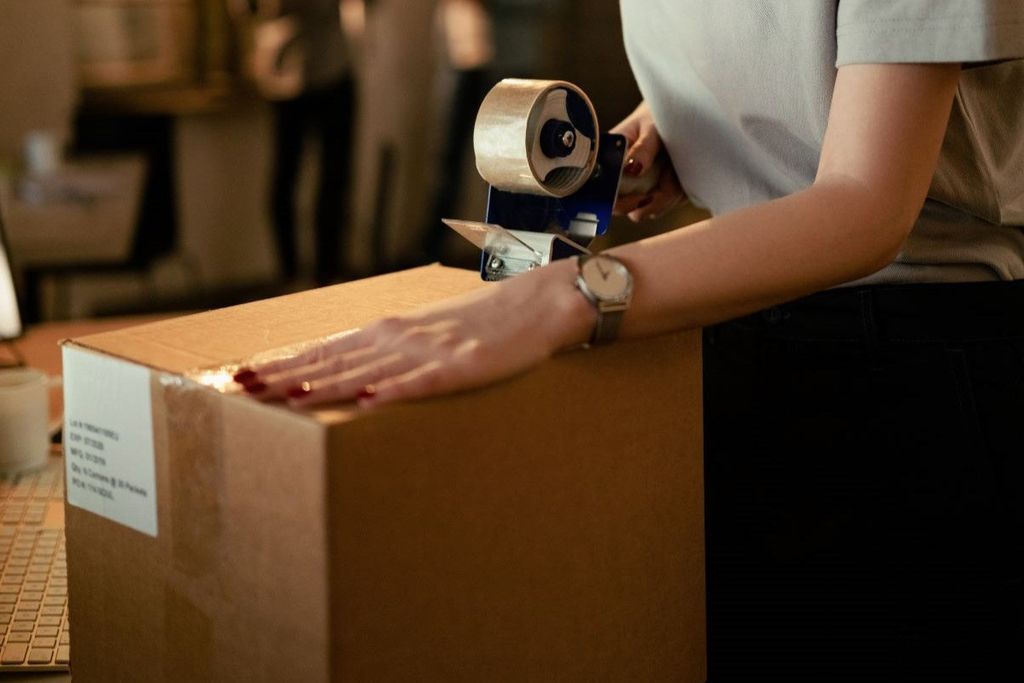 Mujer presiona con la mano la cinta recién aplicada sobre una caja de cartón etiquetada, con dispensador azul