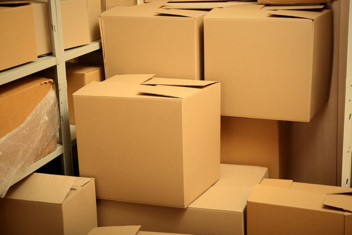Cajas de cartón corrugado de varios tamaños apiladas junto a una estantería metálica en bodega