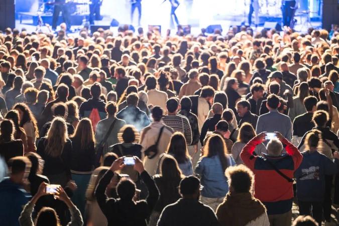 Multitud de espaldas en un concierto al aire libre, con personas grabando con el celular frente al escenario iluminado
