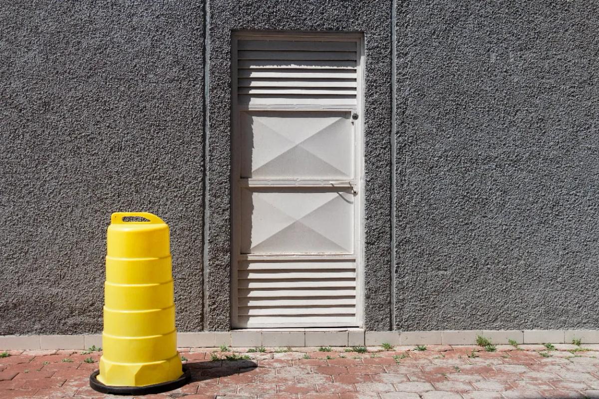 Trafitambo amarillo con asa superior sobre base negra, junto a un muro gris y una puerta blanca en una banqueta de adoquín