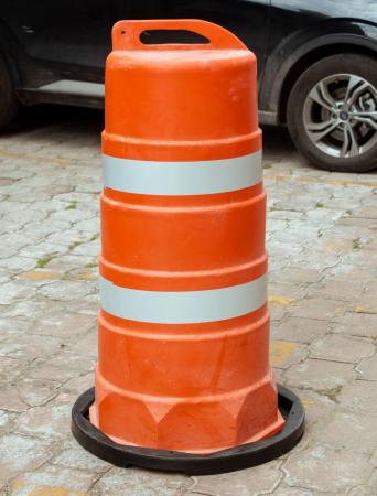 Trafitambo naranja con dos bandas reflejantes blancas y base negra sobre adoquín, con un auto negro estacionado detrás