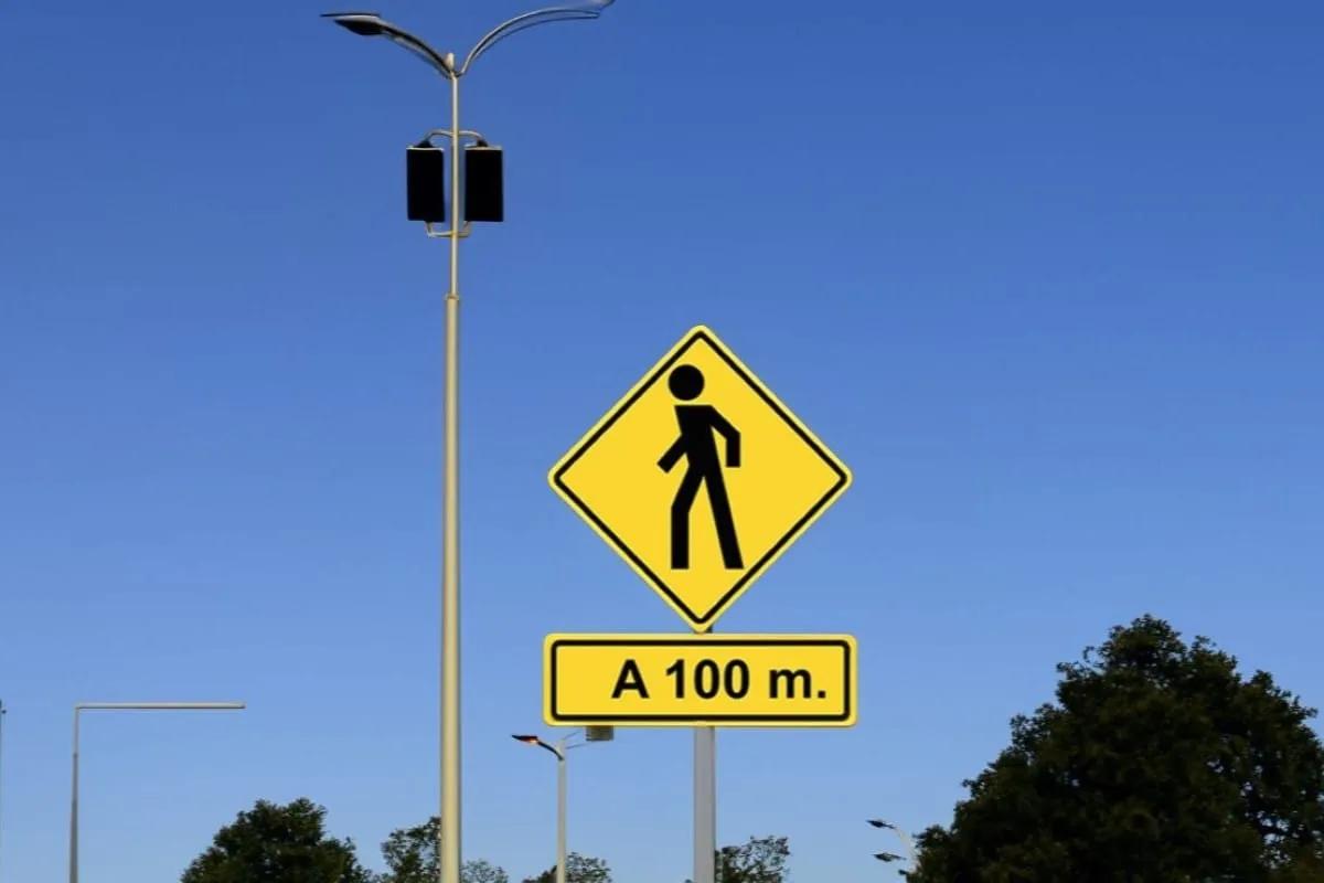 Señal preventiva amarilla de cruce de peatones con placa «A 100 m.» en un poste, con cielo azul