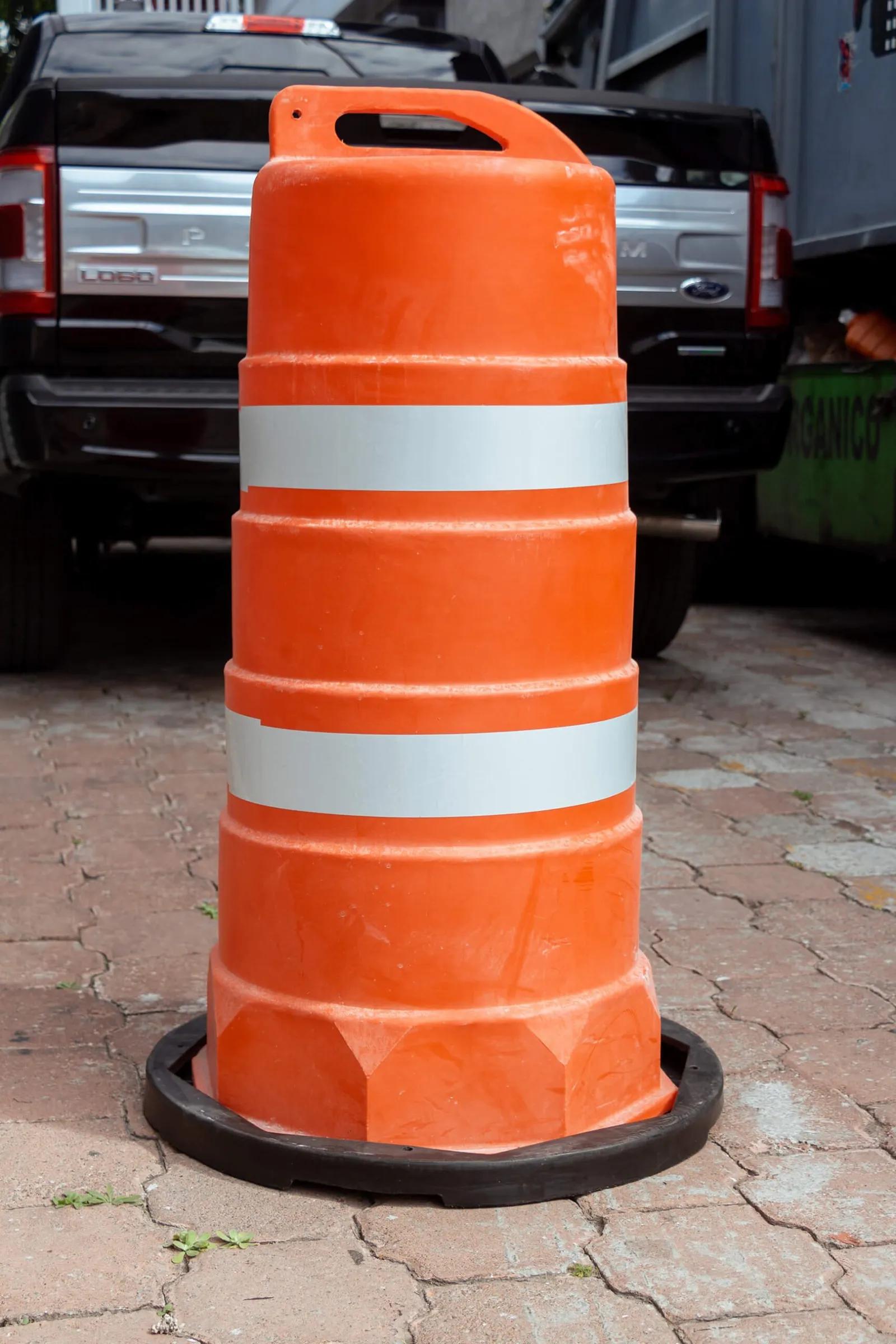 Trafitambo naranja con dos bandas reflejantes blancas sobre base negra en piso de adoquín, con una camioneta negra detrás