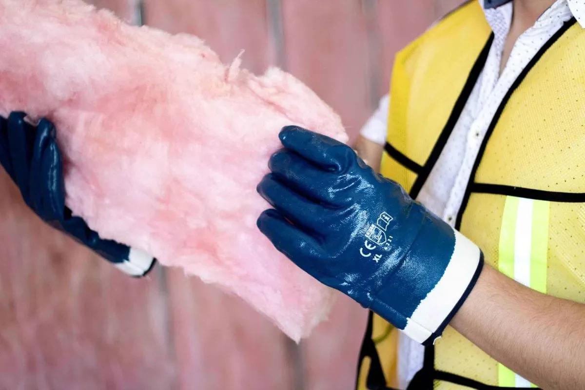 Trabajador con chaleco amarillo y guantes de nitrilo azul sostiene un trozo de aislante rosa de fibra de vidrio