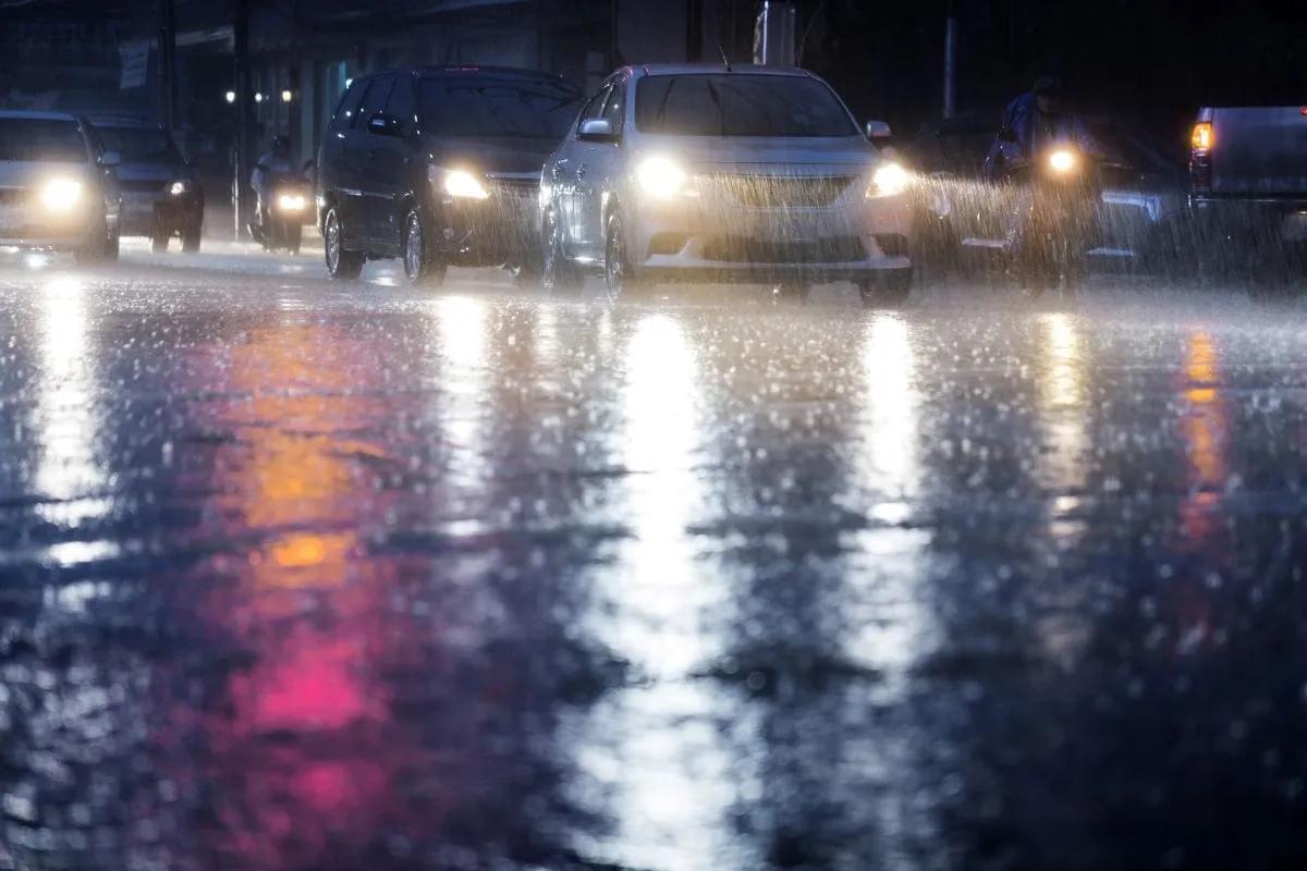 Autos con los faros encendidos bajo lluvia intensa de noche sobre asfalto mojado que refleja las luces