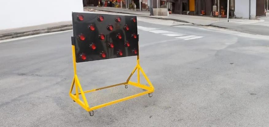 Flechero luminoso de panel negro con luces rojas sobre un soporte amarillo con ruedas en una calle
