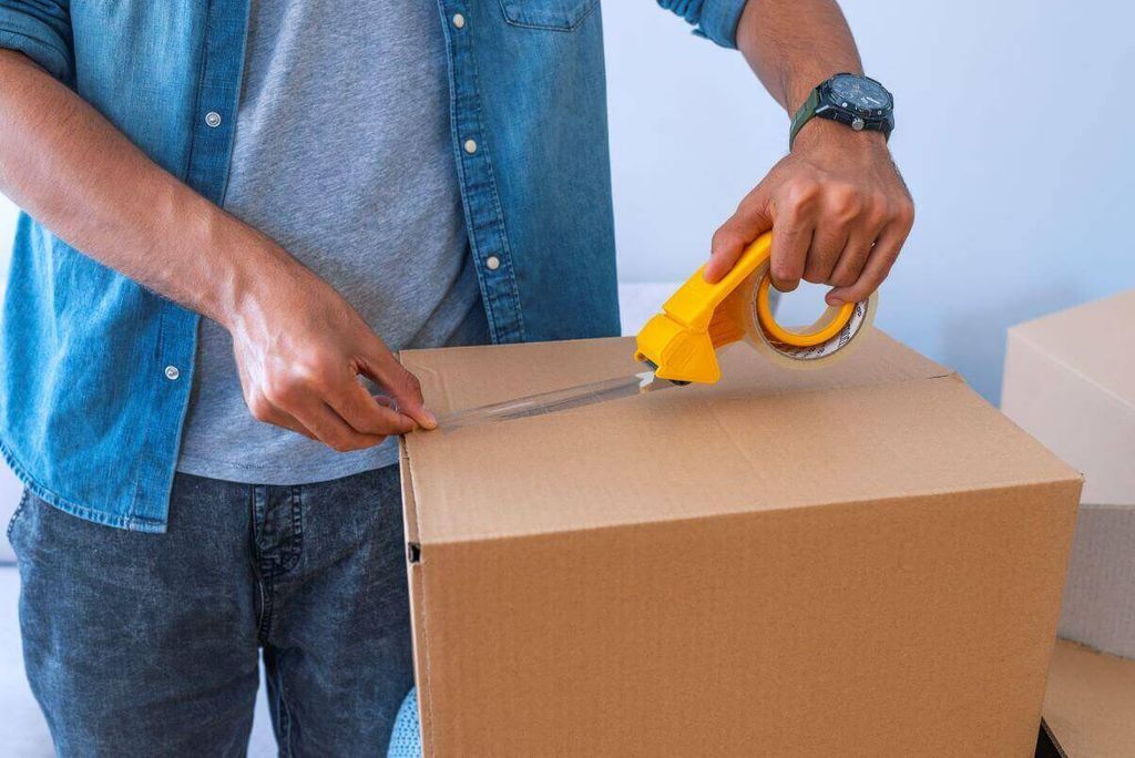 Hombre con camisa de mezclilla sella una caja de cartón con un despachador de cinta transparente