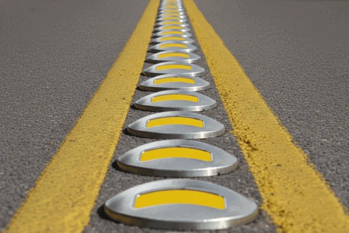 Fila de botones viales metálicos con reflejante amarillo entre dos líneas amarillas pintadas sobre el asfalto