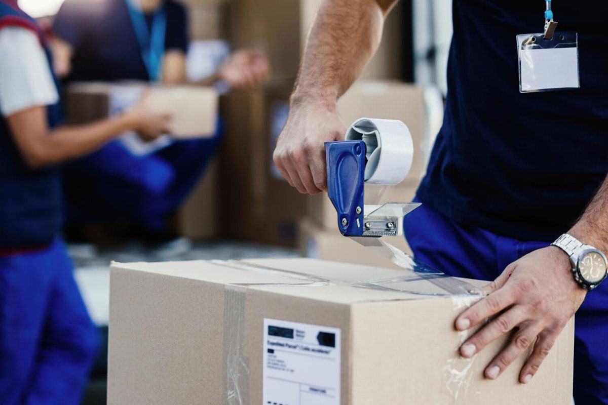 Trabajador con uniforme azul sella una caja de cartón con un dispensador de cinta transparente en una bodega
