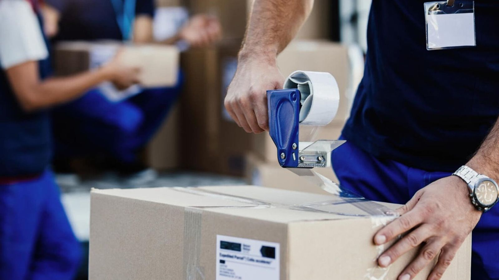 Trabajador con uniforme azul sella una caja de cartón con un dispensador de cinta transparente en una bodega