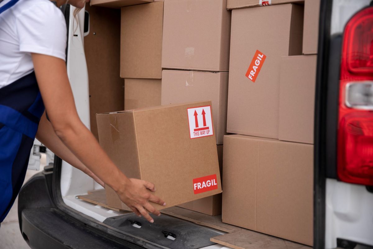 Repartidora carga una caja de cartón con etiquetas de frágil y flechas hacia arriba en una van llena de cajas