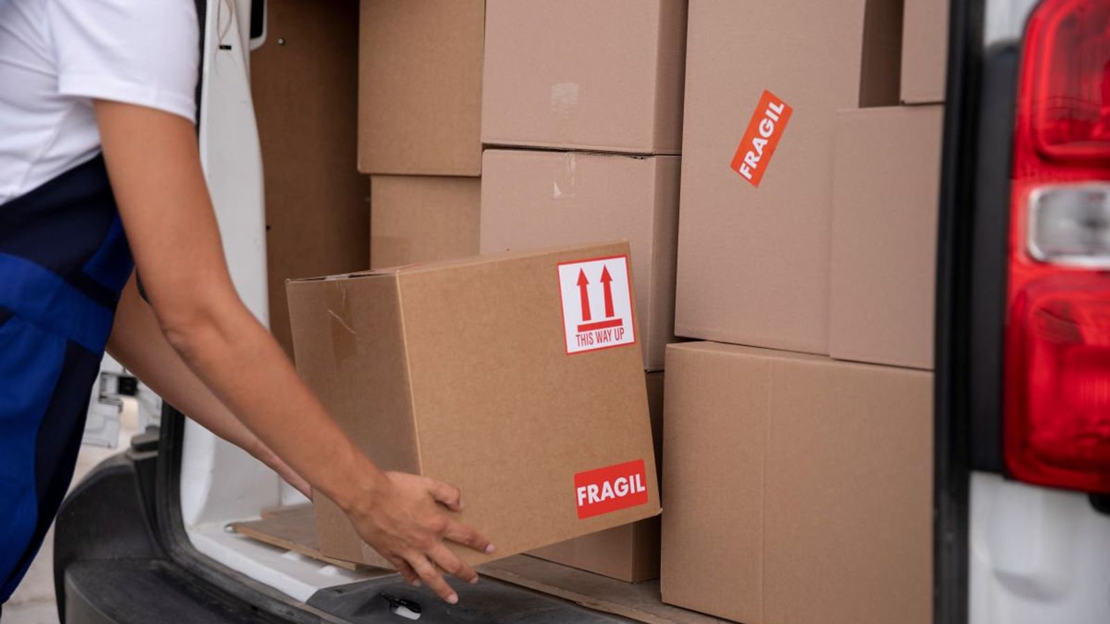 Repartidora carga una caja de cartón con etiquetas de frágil y flechas hacia arriba en una van llena de cajas