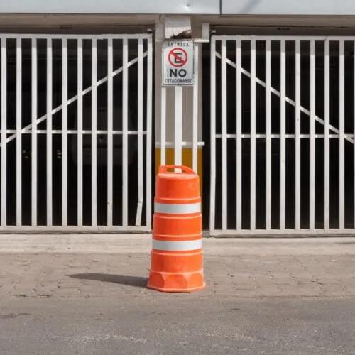 Trafitambo naranja con dos bandas reflejantes frente a un portón blanco con letrero de no estacionarse