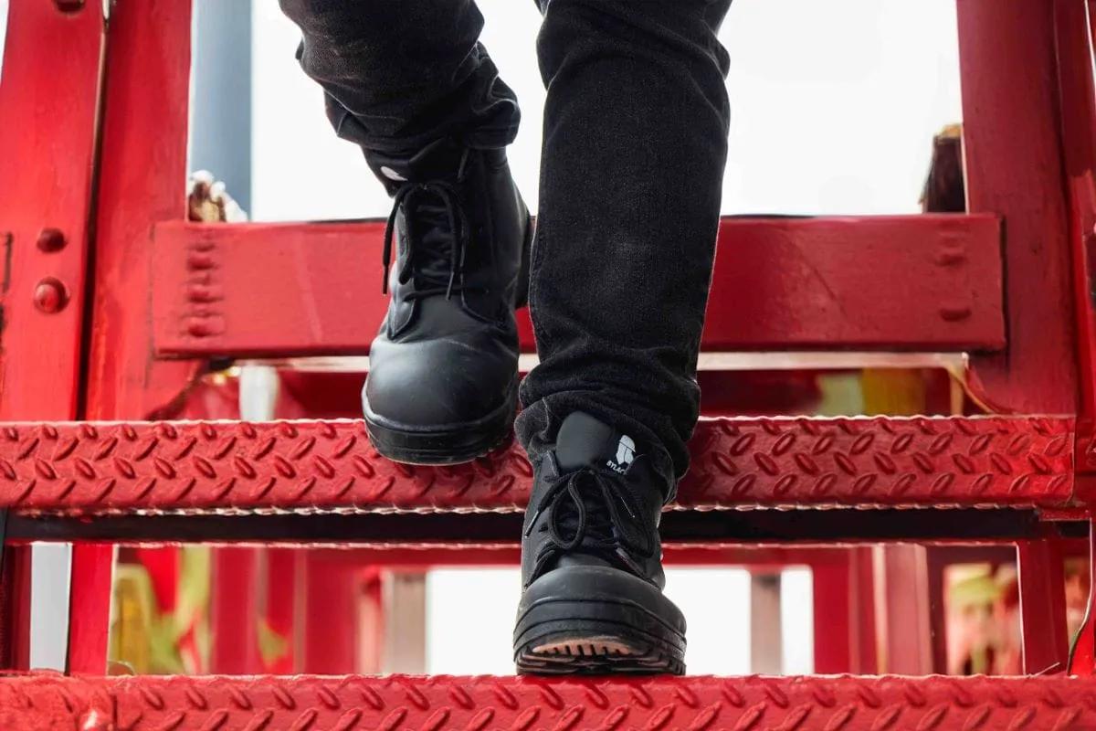 Botas de seguridad negras suben una escalera metálica roja con peldaños antiderrapantes