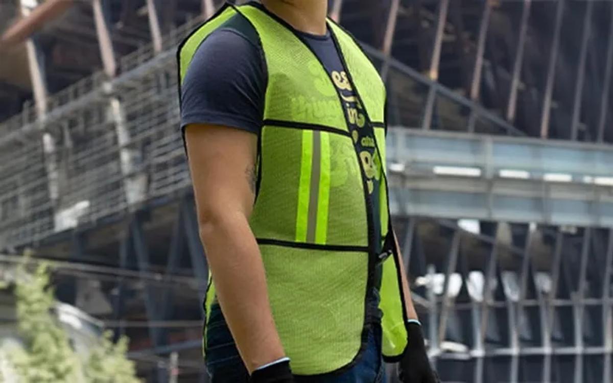 Trabajador con chaleco de malla amarillo neón con franjas reflejantes y guantes negros frente a una estructura de acero