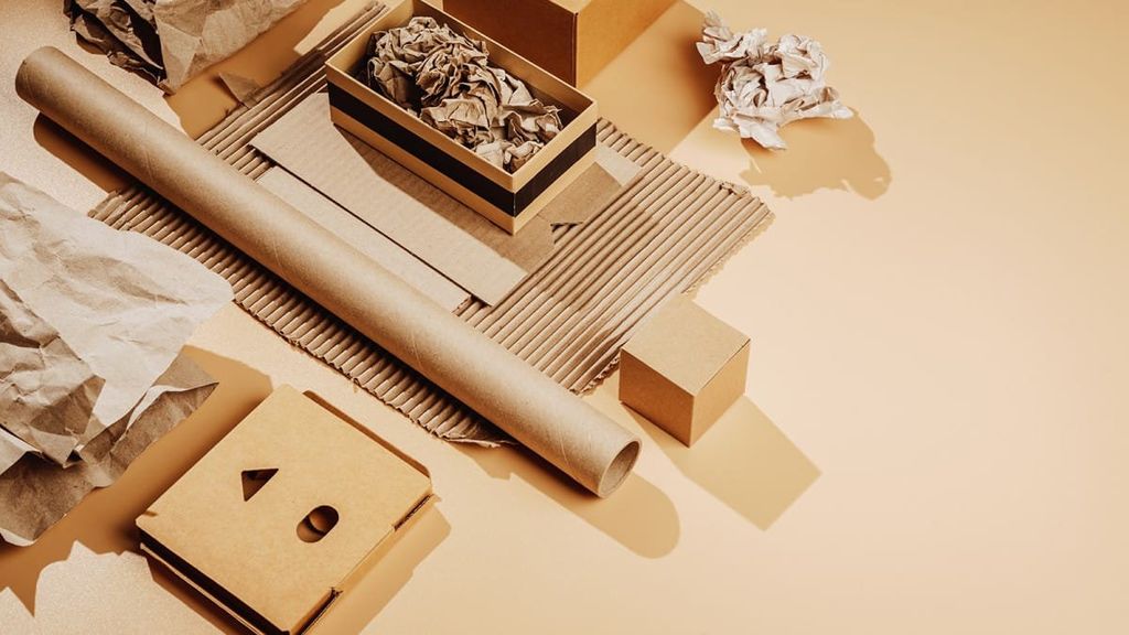 Materiales de cartón sobre fondo beige: tubo, corrugado, caja con papel arrugado y esquineros troquelados