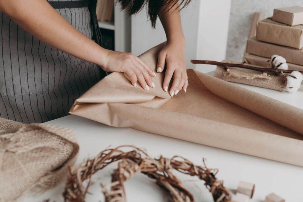 Persona con mandil a rayas dobla papel kraft sobre una mesa blanca, con cajas kraft al fondo