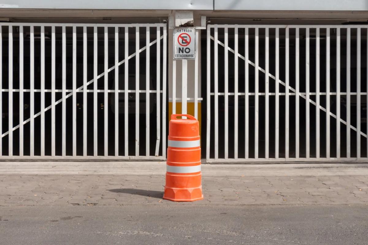 Trafitambo naranja con dos bandas reflejantes frente a un portón blanco con letrero de no estacionarse