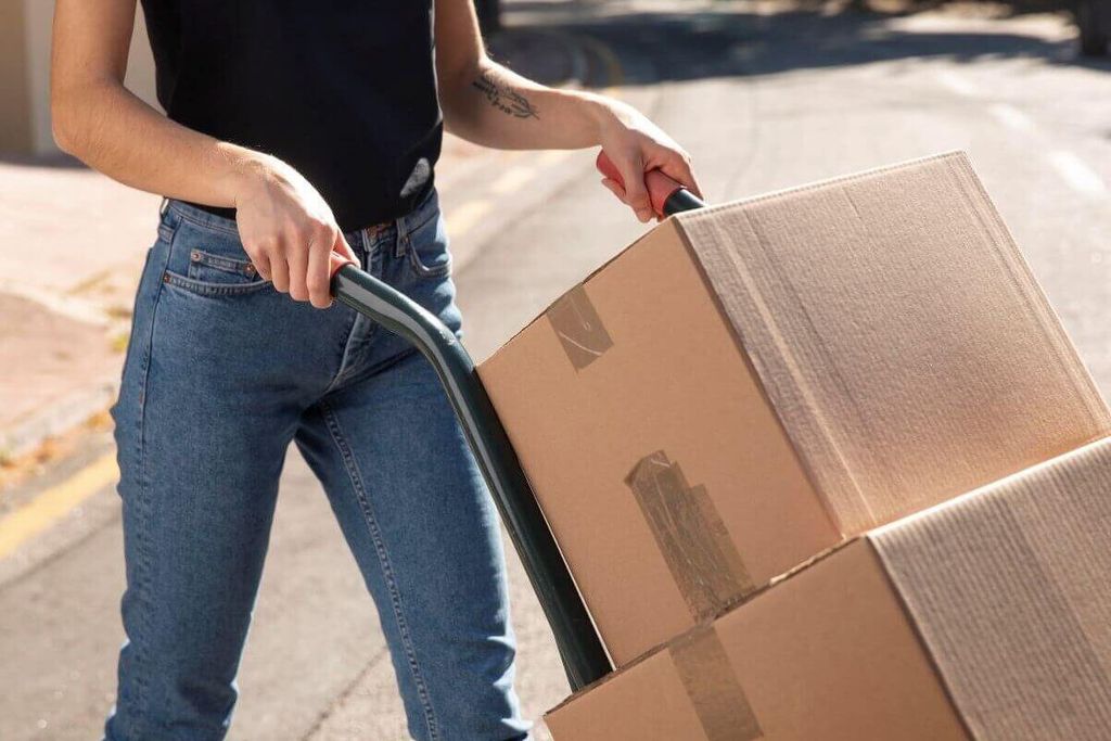 Persona en jeans empuja un diablito con dos cajas de cartón selladas con cinta por una banqueta