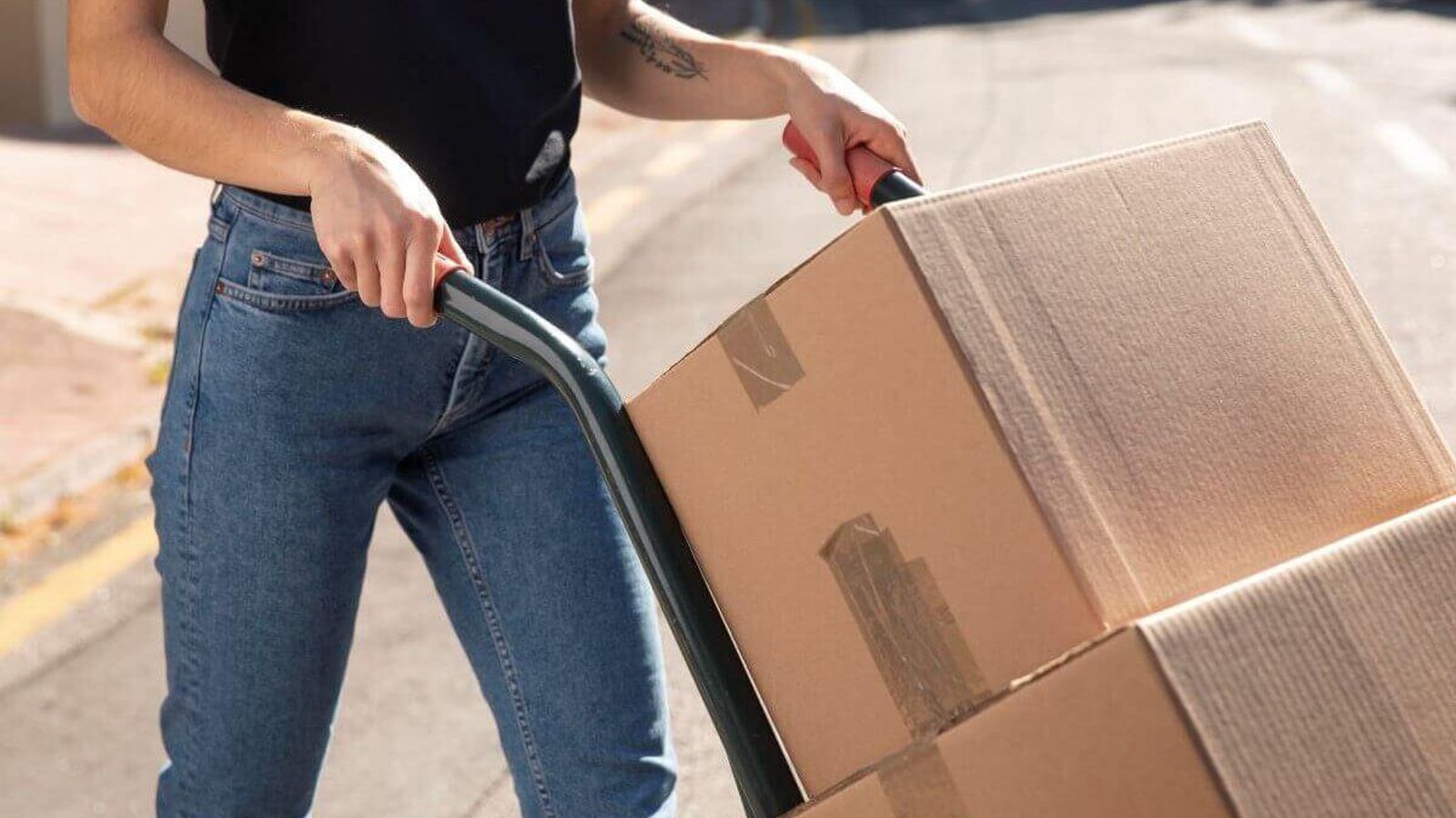 Persona empuja un diablito con dos cajas de cartón selladas con cinta por una calle soleada