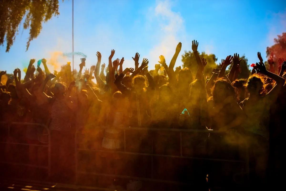 Público con los brazos en alto entre nubes de polvo de colores durante un festival, detrás de vallas metálicas