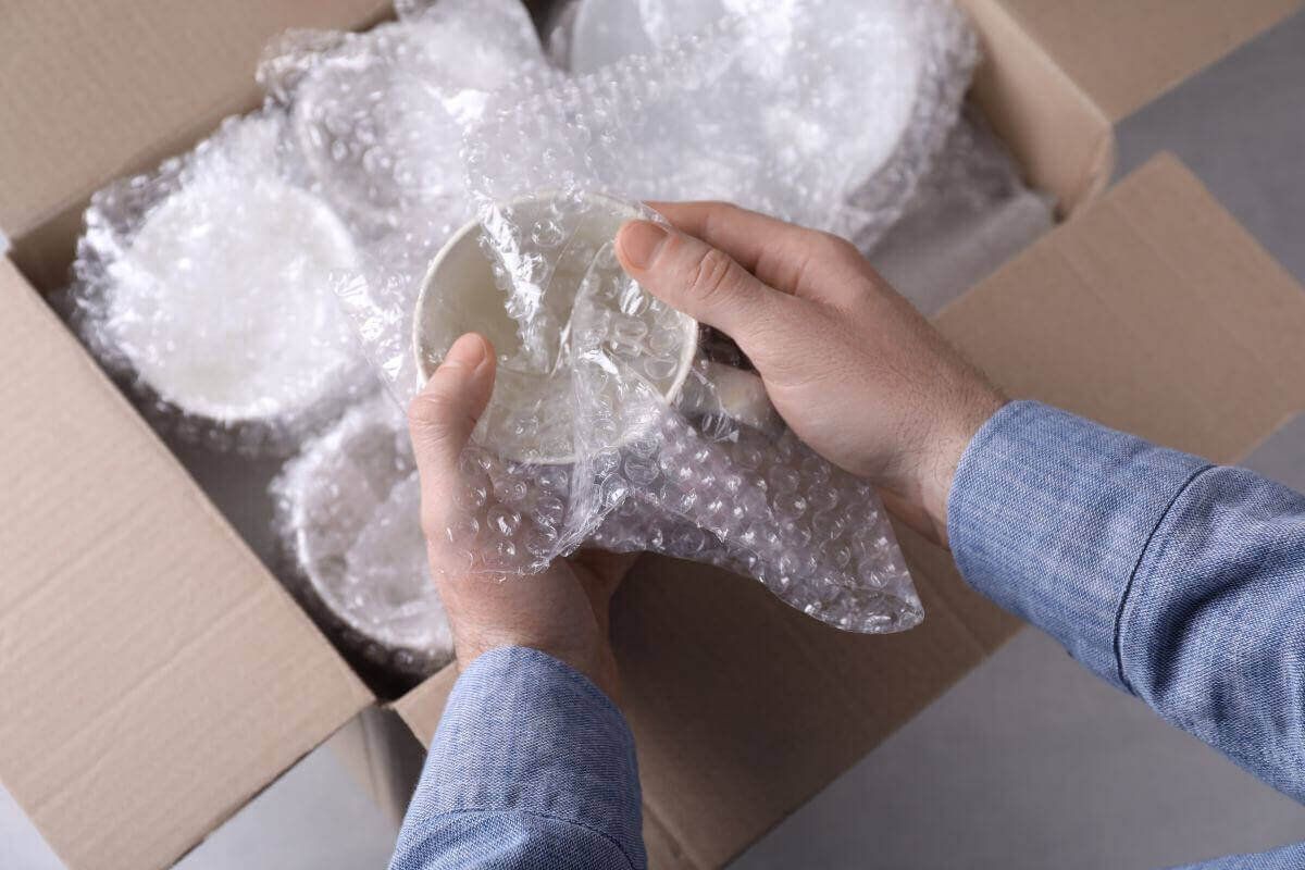 Manos envuelven un tazón de cerámica en poliburbuja sobre una caja de cartón con más piezas ya envueltas
